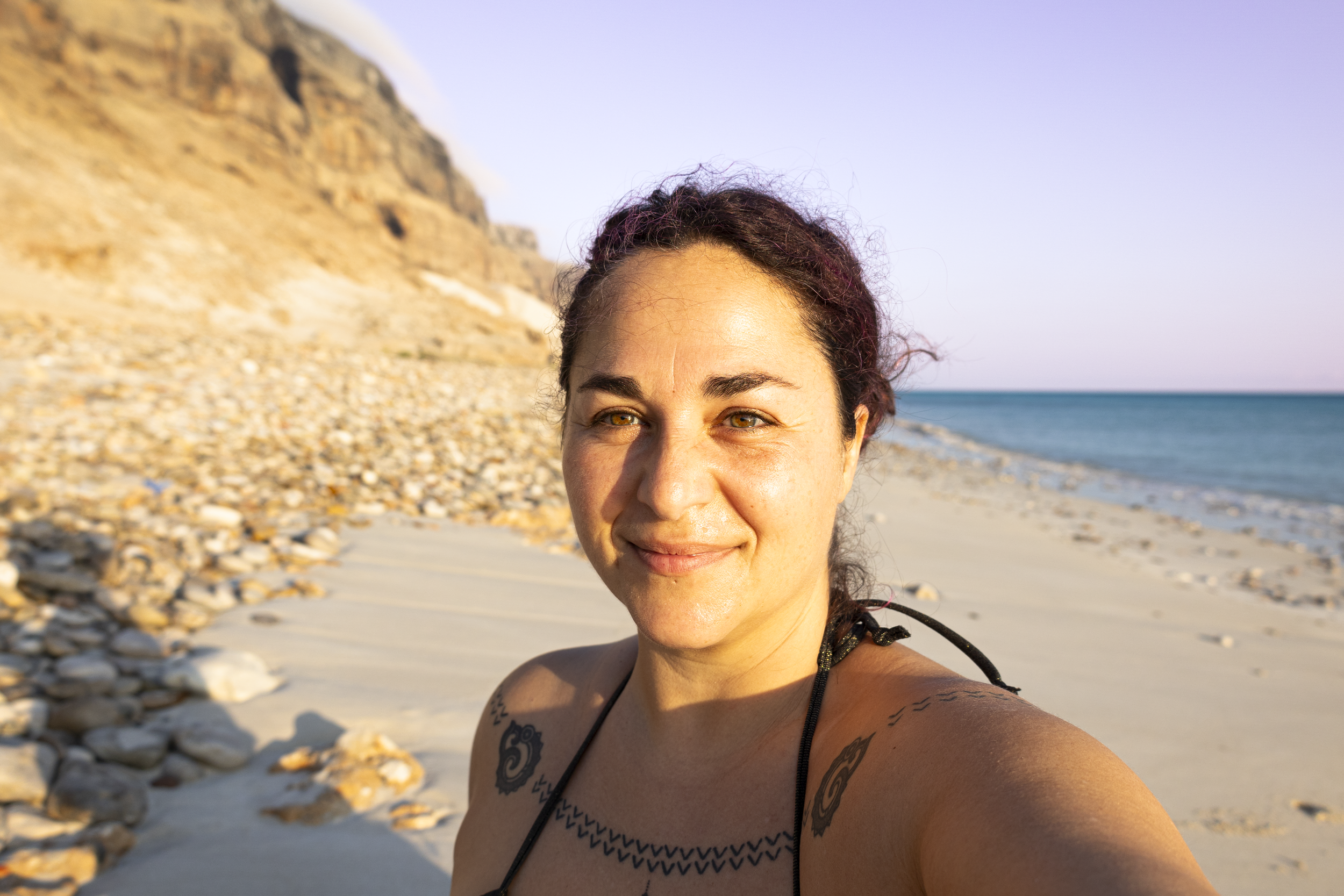 Francesca, a woman with tattoos smiling at the camera on a beach on Socotra Island, Yemen, with rocks and cliffs in the background.