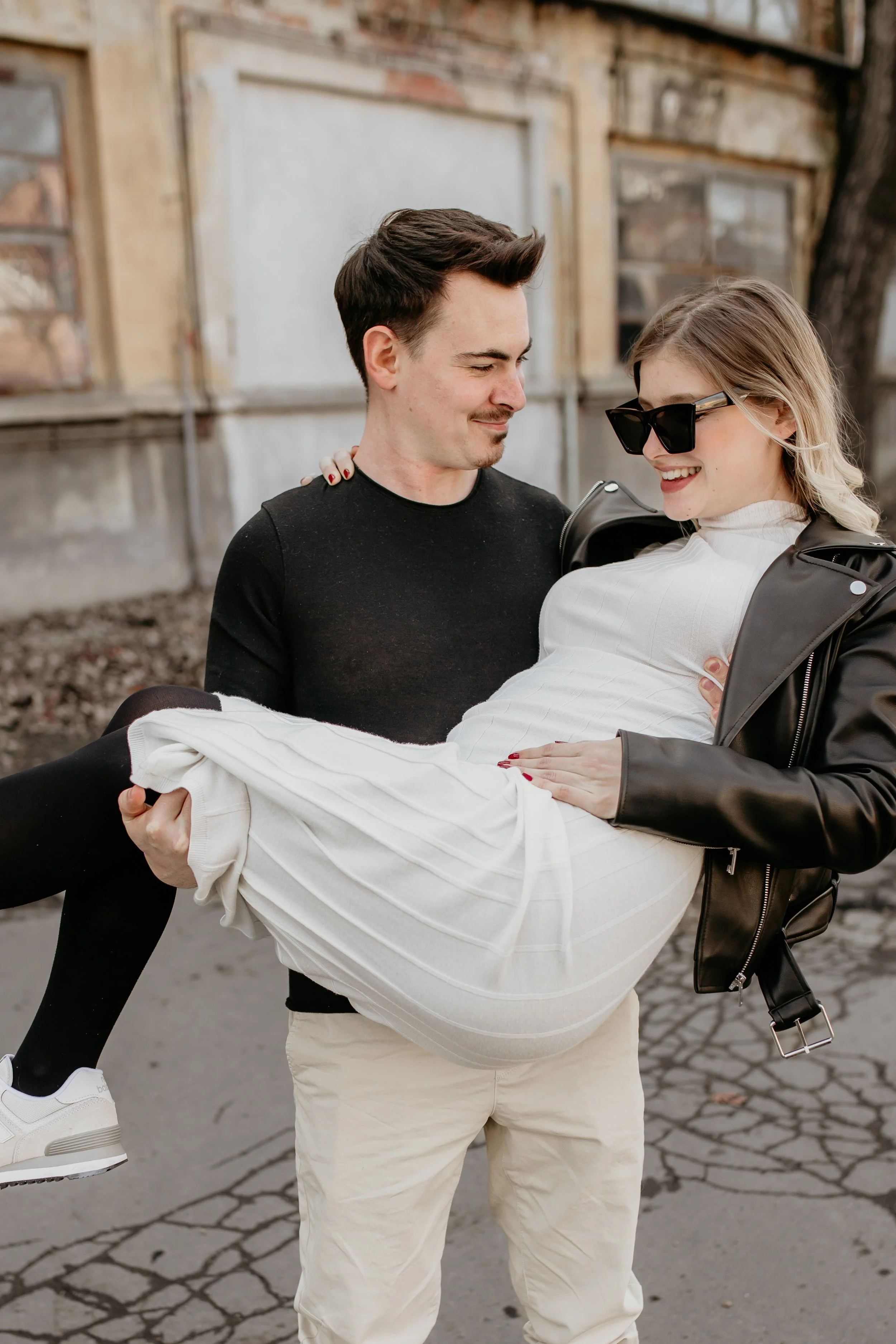 A man holding a woman in his arms outdoors, with the woman smiling and wearing sunglasses, against a background of an old building with a tree nearby.