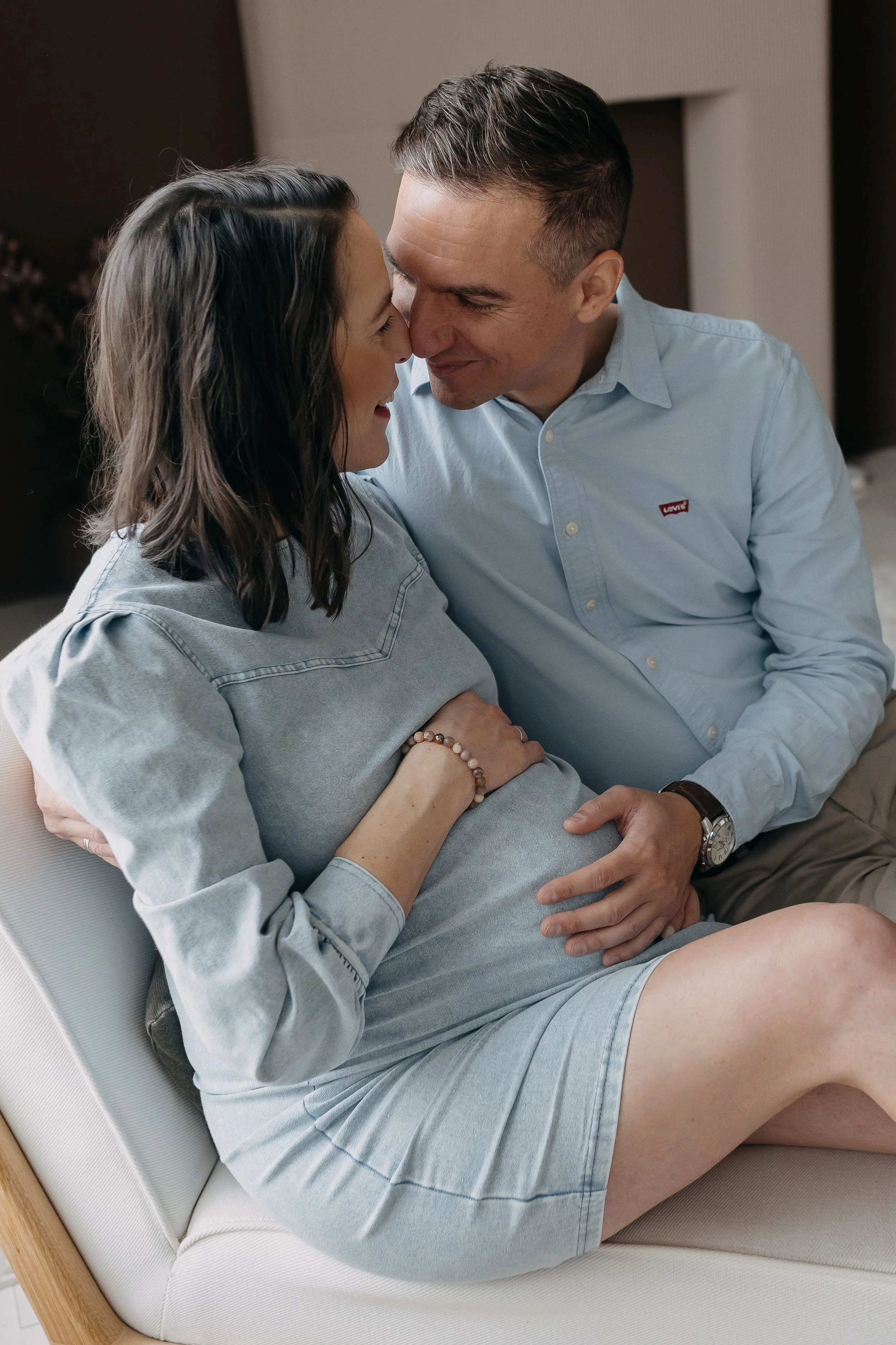 A man and woman are sitting close together on a white sofa, with heads touching, smiling and sharing an intimate moment.