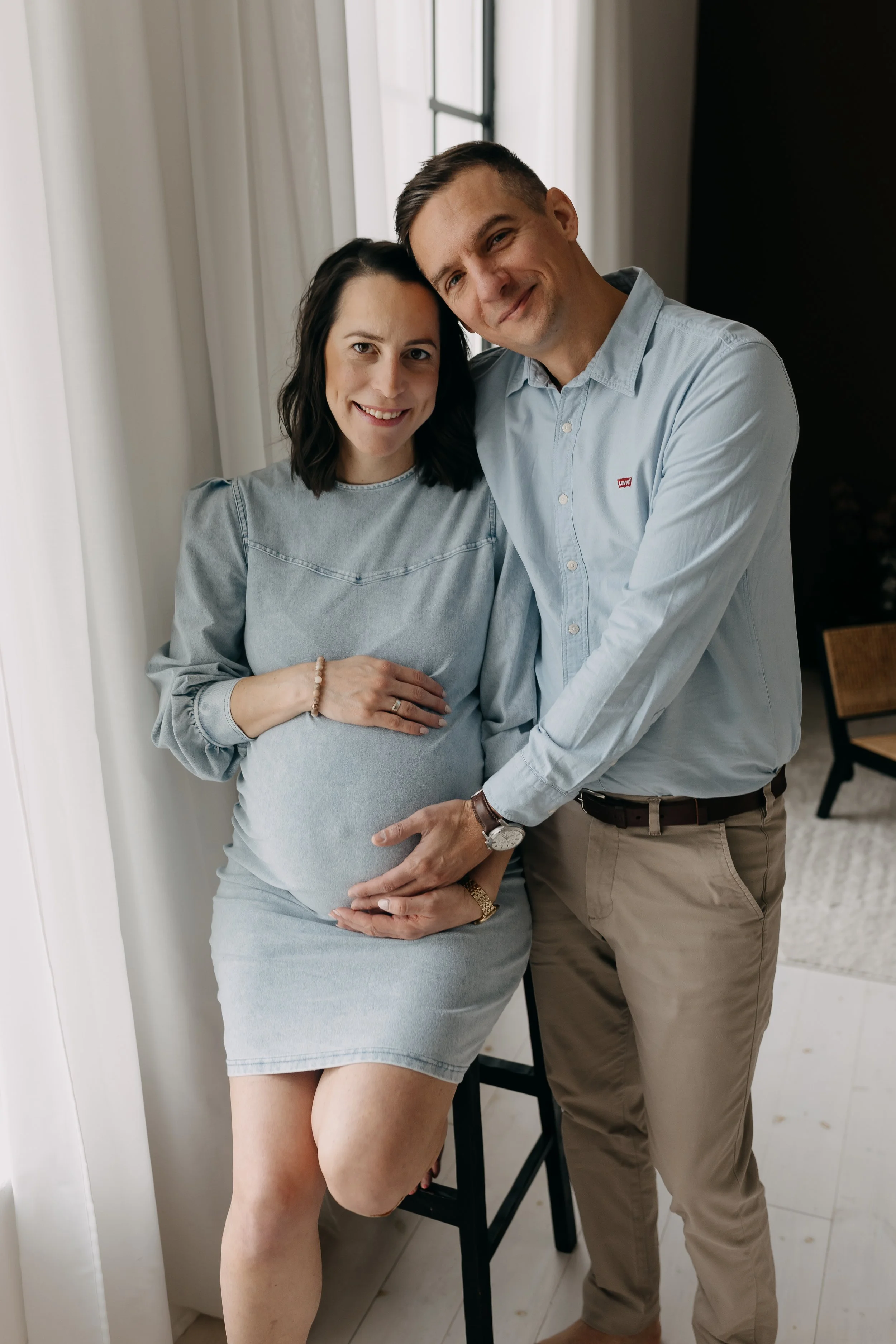 A pregnant woman and a man are standing close together indoors near a window. The woman has dark hair and is wearing a light gray dress, smiling and holding her belly. The man has short brown hair, is wearing a light blue button-up shirt with a small