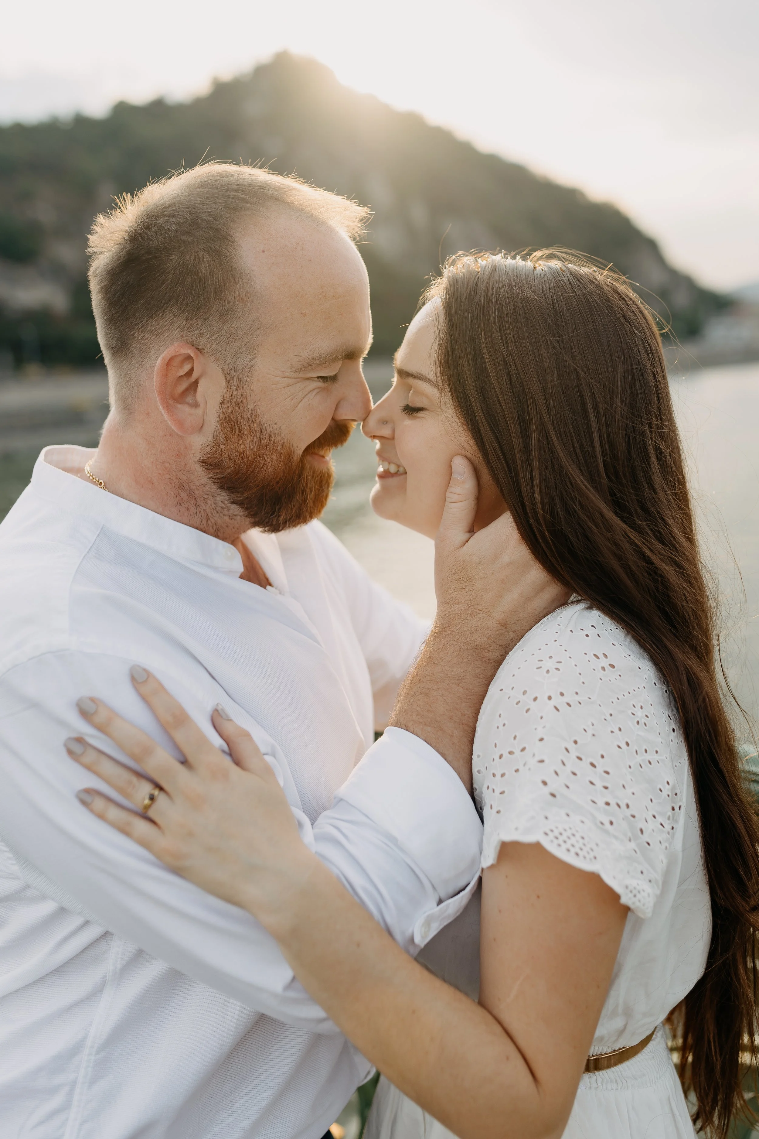 A man and woman share an intimate moment outdoors, touching foreheads and smiling with eyes closed, with a mountain and water in the background during sunset.