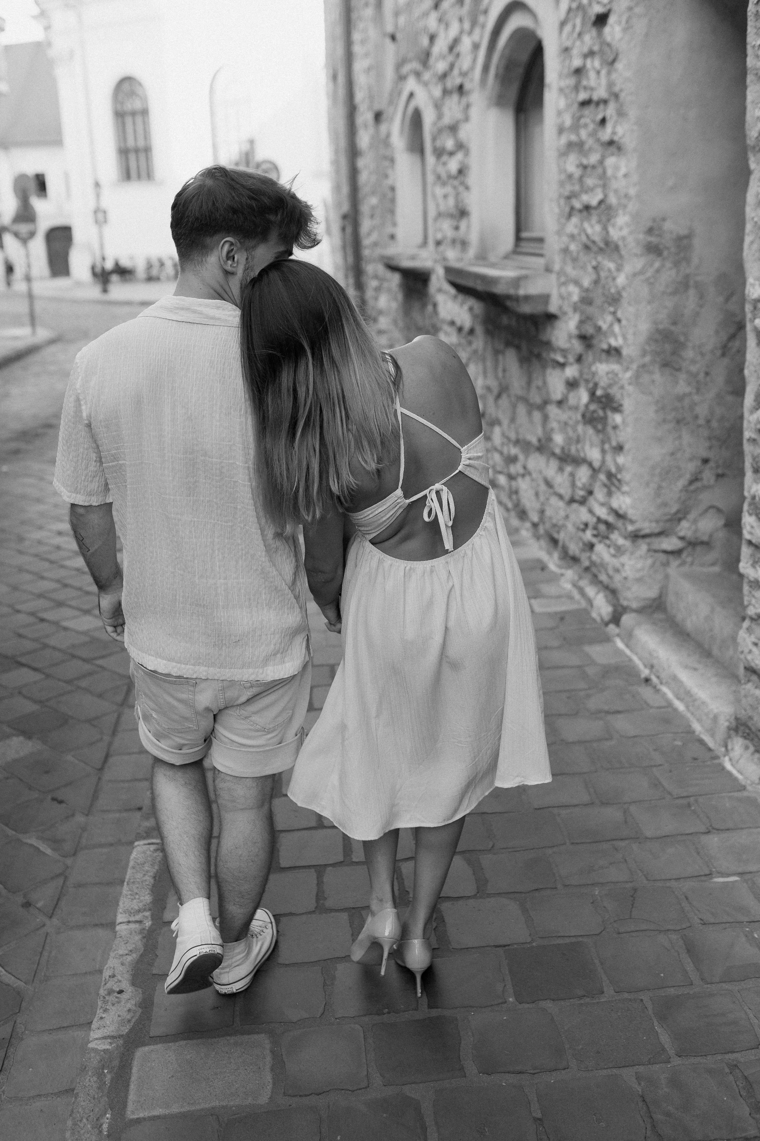 A black and white photo of a young couple walking closely together on a cobblestone street, with the man's head leaning on the woman's shoulder.