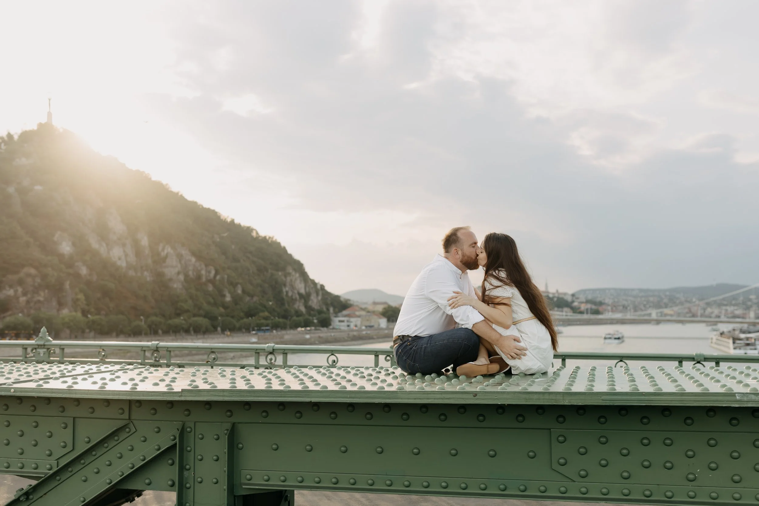 A couple sits close together on a green bridge, sharing a kiss with a river, a city skyline, and a hill with a monument in the background during daytime.