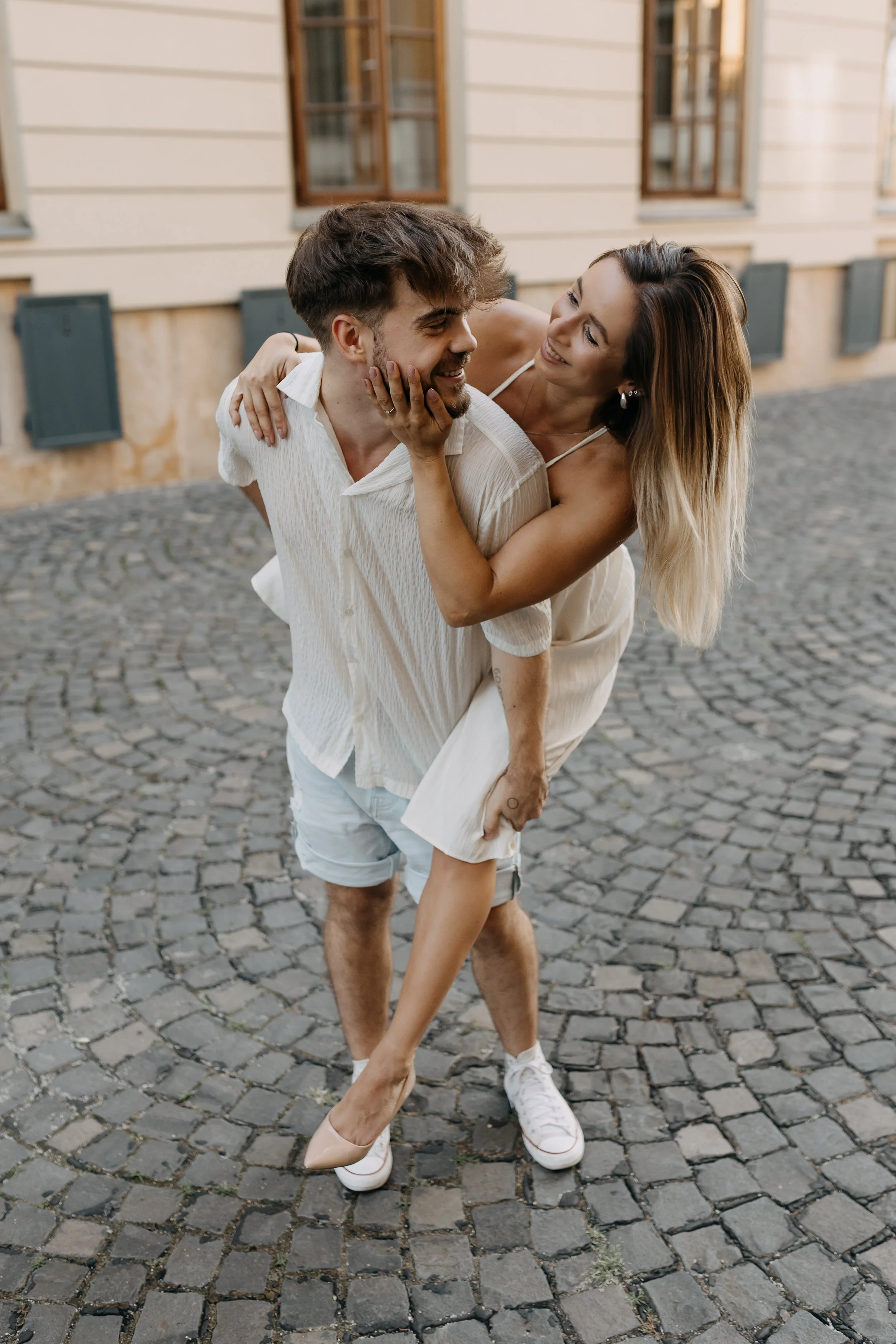 A couple sharing a playful moment outdoors on a cobblestone street, with the woman giving the man a piggyback ride, smiling at each other.