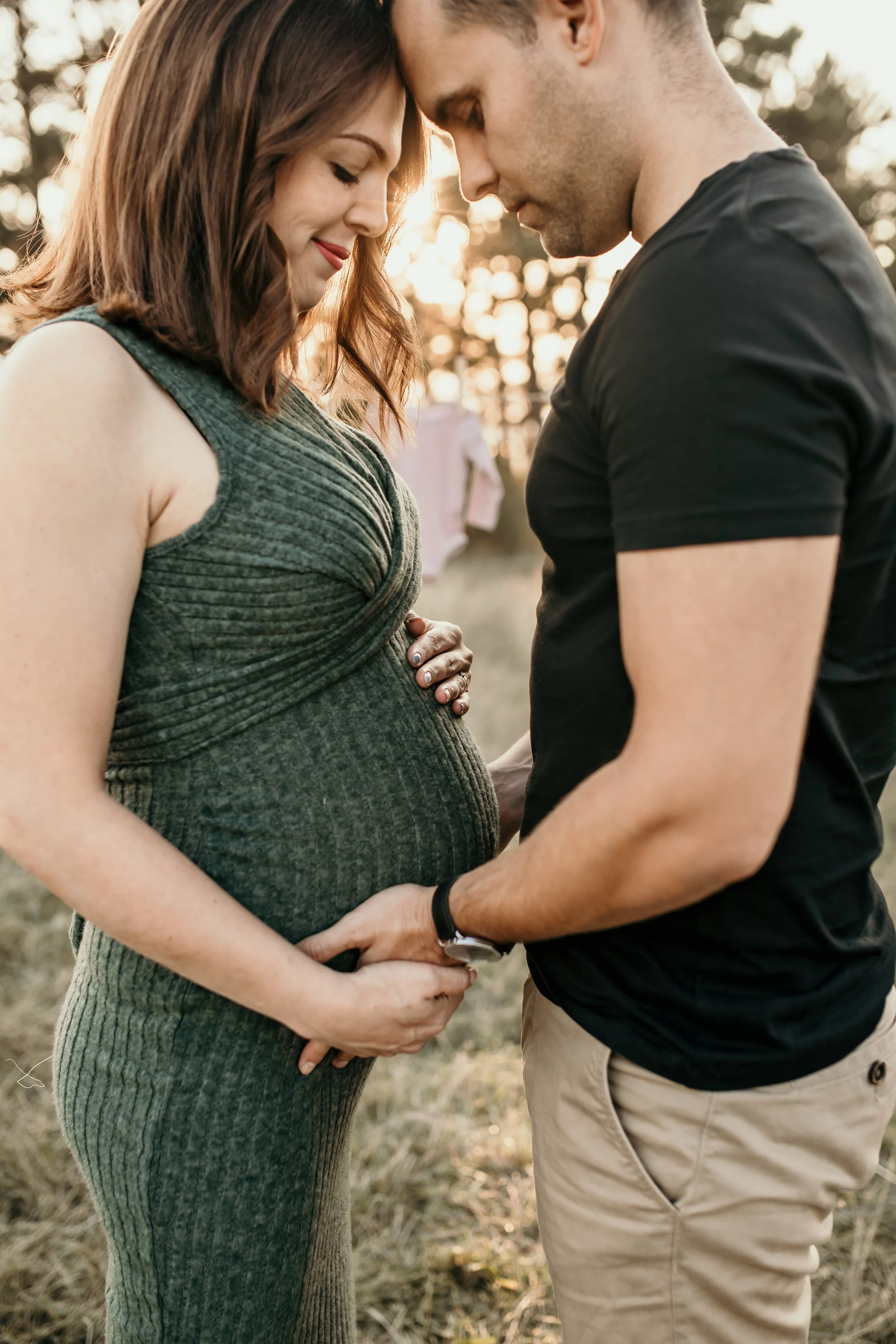A pregnant woman and a man are standing close together outdoors at sunset, gently holding her belly with their foreheads touching and eyes closed, sharing an intimate moment.