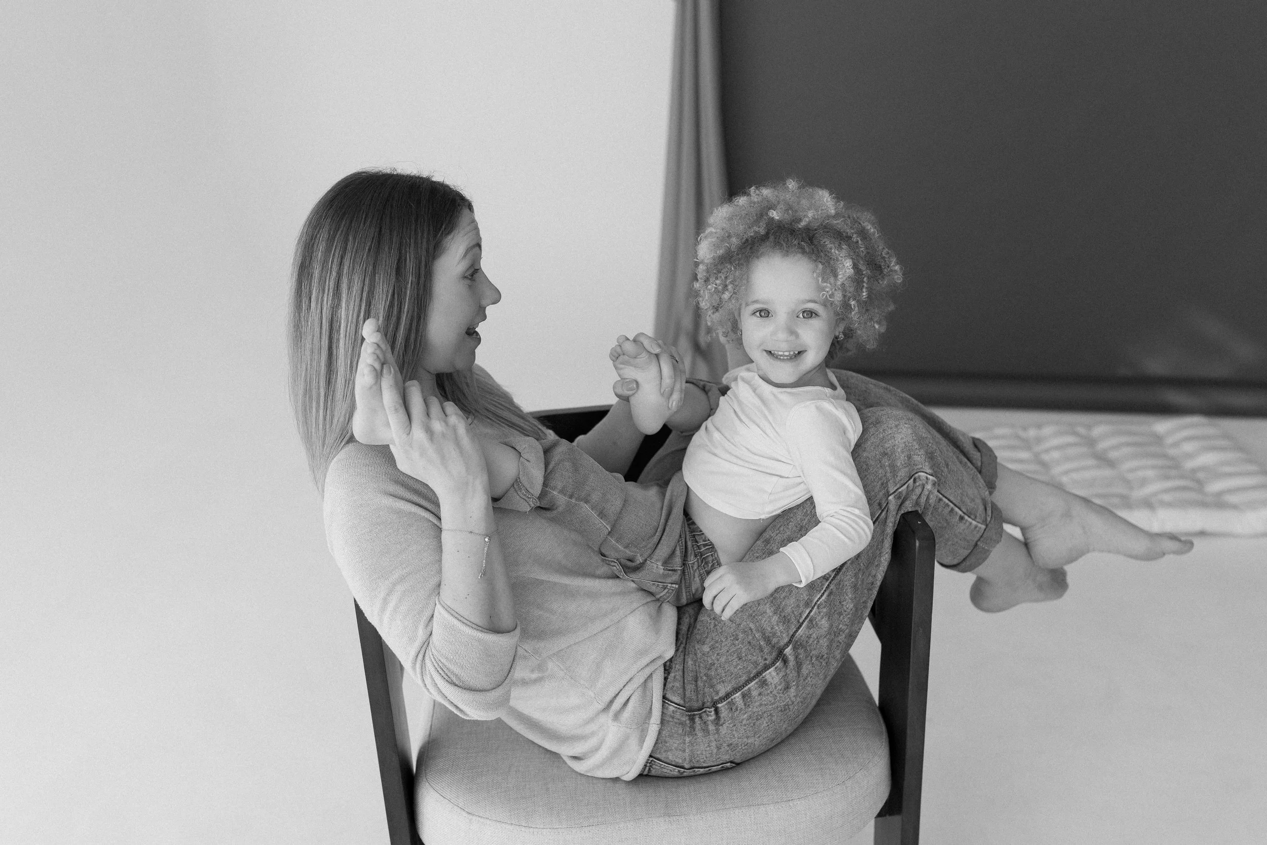 A woman and a young girl with curly hair playing together indoors, smiling, with the girl sitting on the woman's lap and holding her hand.