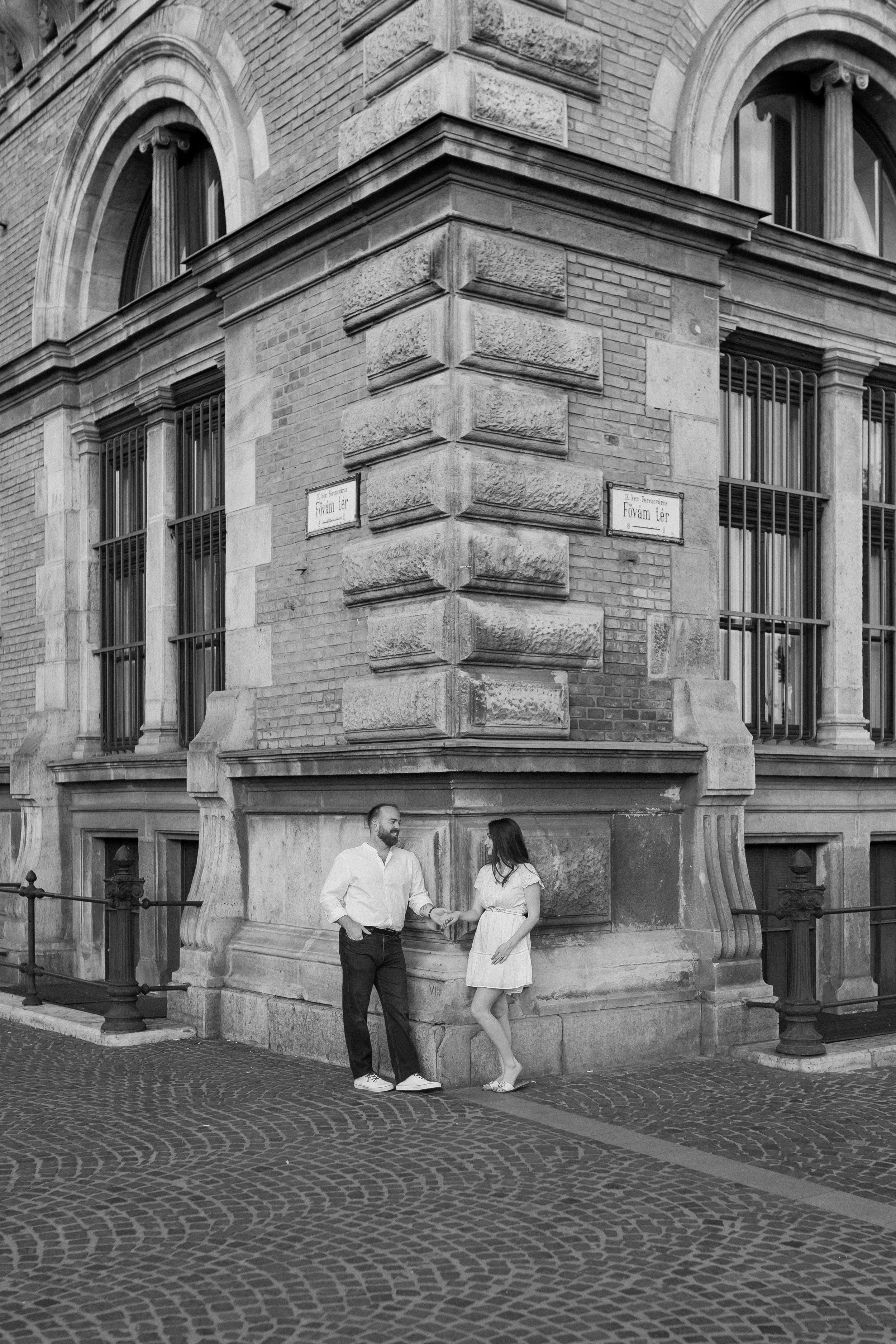 A black-and-white photo of a man and woman standing on a curved cobblestone street outside a historic brick building with arched windows and decorative stone details. The man has a beard, is wearing a white shirt and dark pants, and is shaking hands 