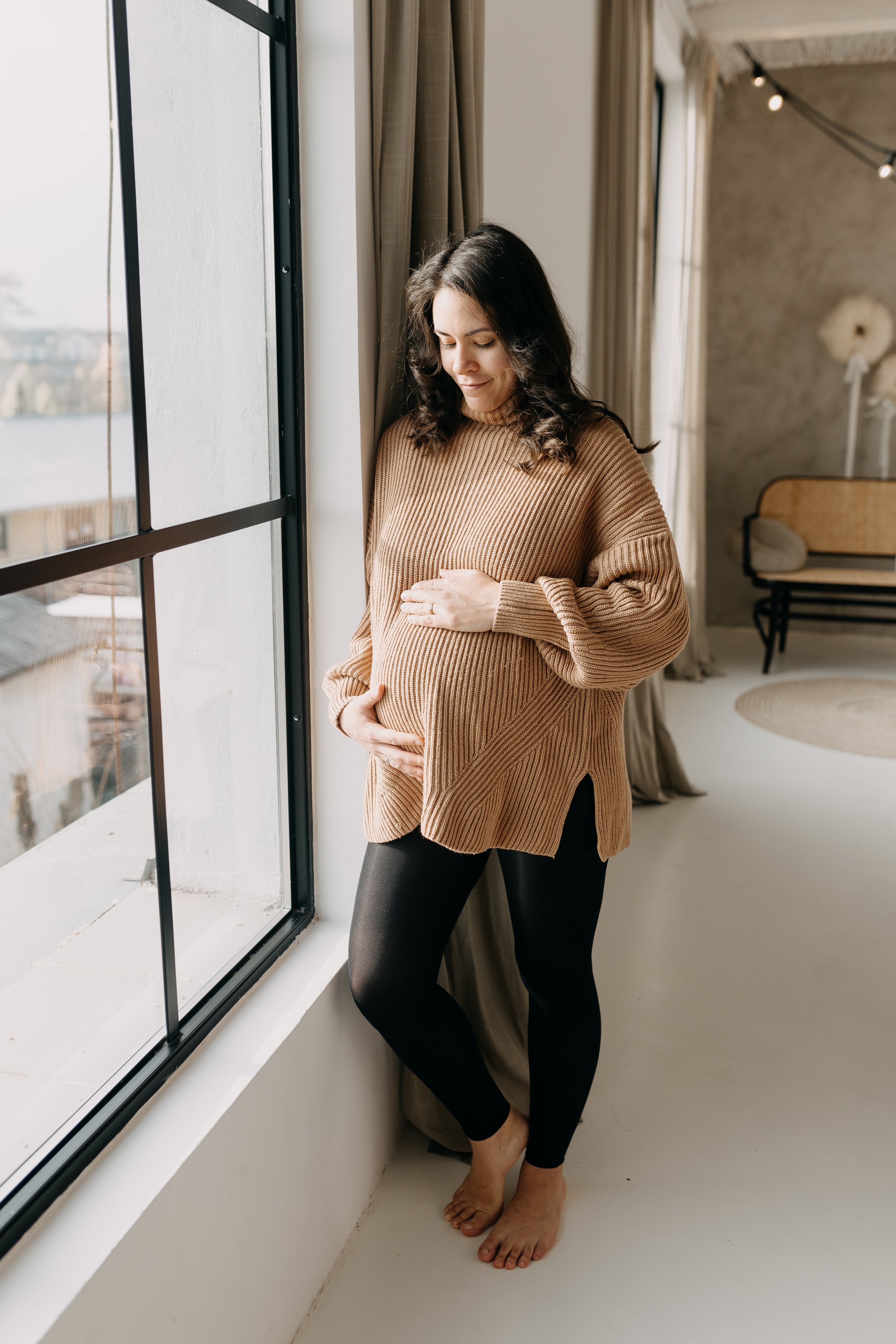 A pregnant woman with dark, curly hair standing by a large window in a modern room. She is wearing a tan, ribbed sweater and black leggings, touching her belly with one hand and resting the other on her chest. The room has neutral decor, curtains, an