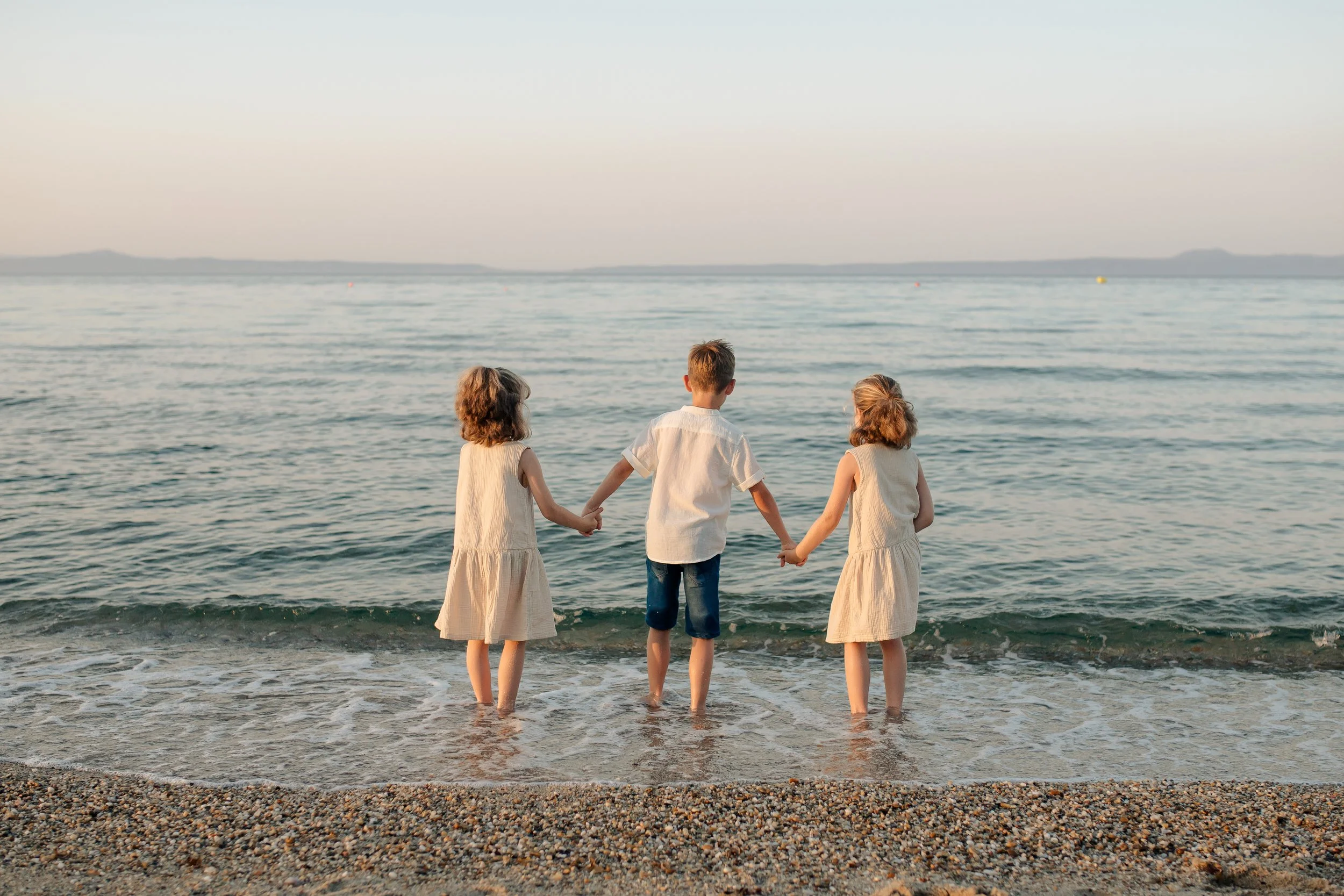 Two children, a boy and a girl, standing on a rocky shoreline by the water. The girl is hugging the boy and leaning in to kiss him on the cheek. The boy is smiling at the camera, and there is a boat in the distance on the water.