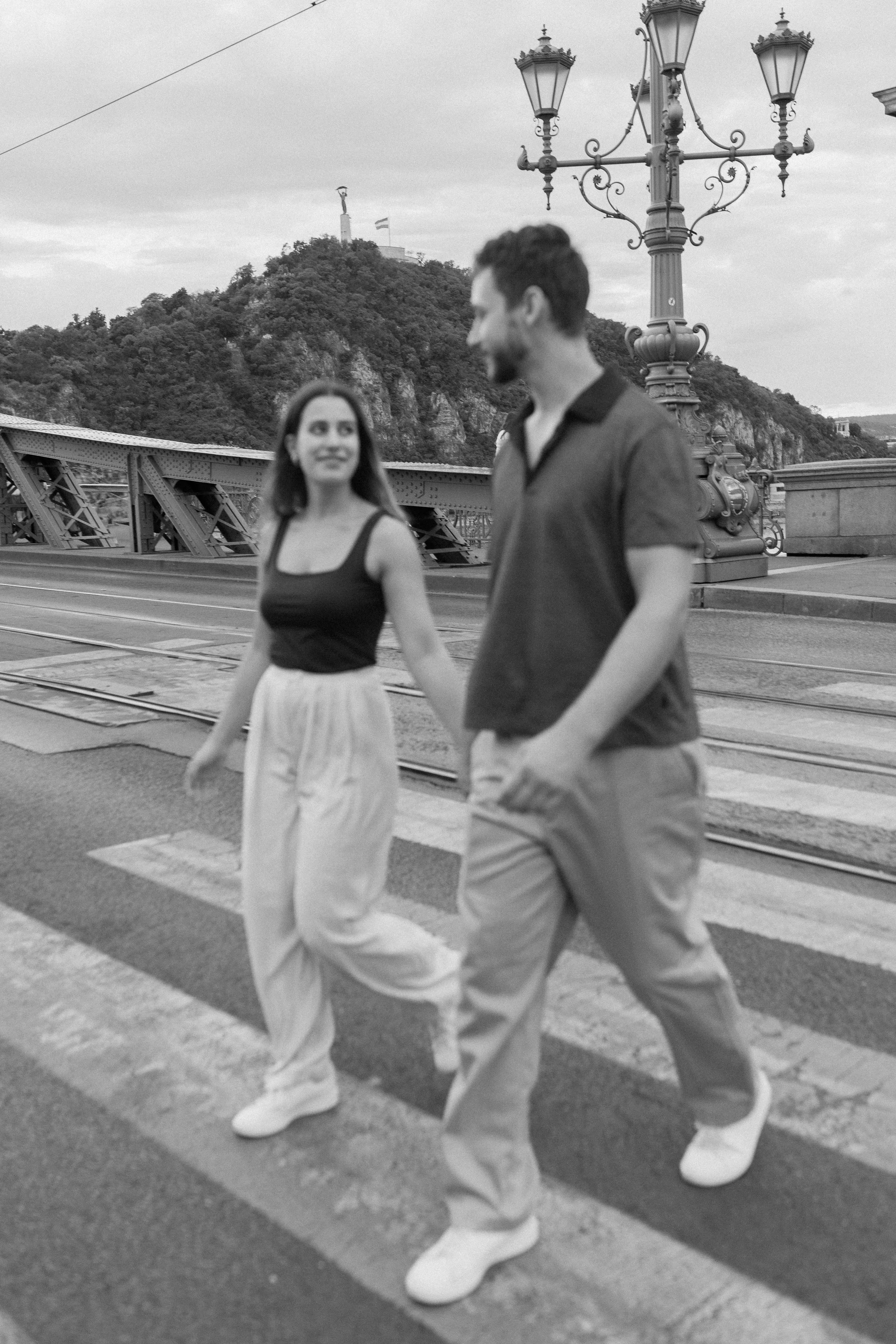 A black and white photo of a young woman and man walking across a street with a pedestrian crossing. The woman is wearing a sleeveless top and a long skirt, looking at the man. The man is wearing a short-sleeved shirt and pants, looking back at the w
