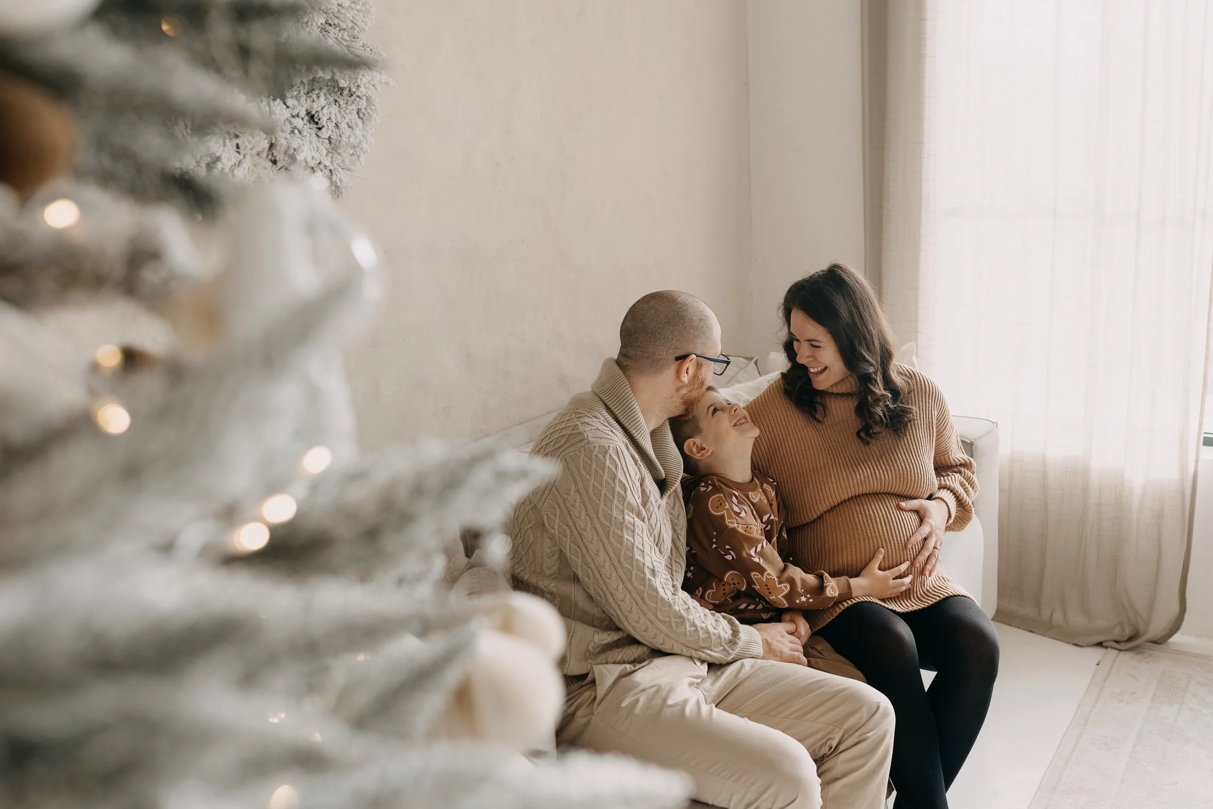 A family sitting on a couch during Christmas, with a decorated white Christmas tree in the foreground. The family includes a pregnant woman, a man, and a young girl, all smiling and looking at each other.