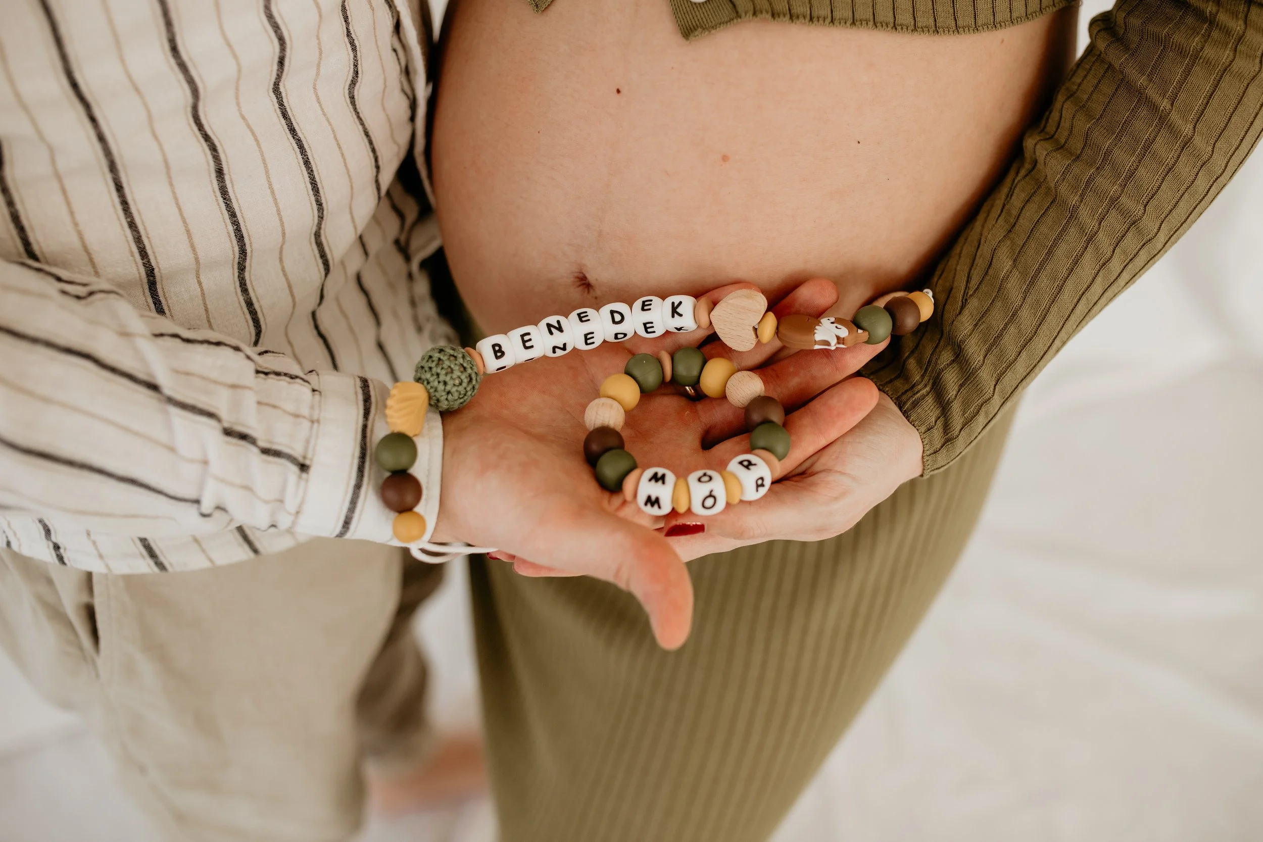 Close-up of a pregnant woman’s bare belly with a person holding a beaded bracelet that spells 'Benedek' in white letter beads, with additional colorful beads, against a background of striped and solid-colored clothing.