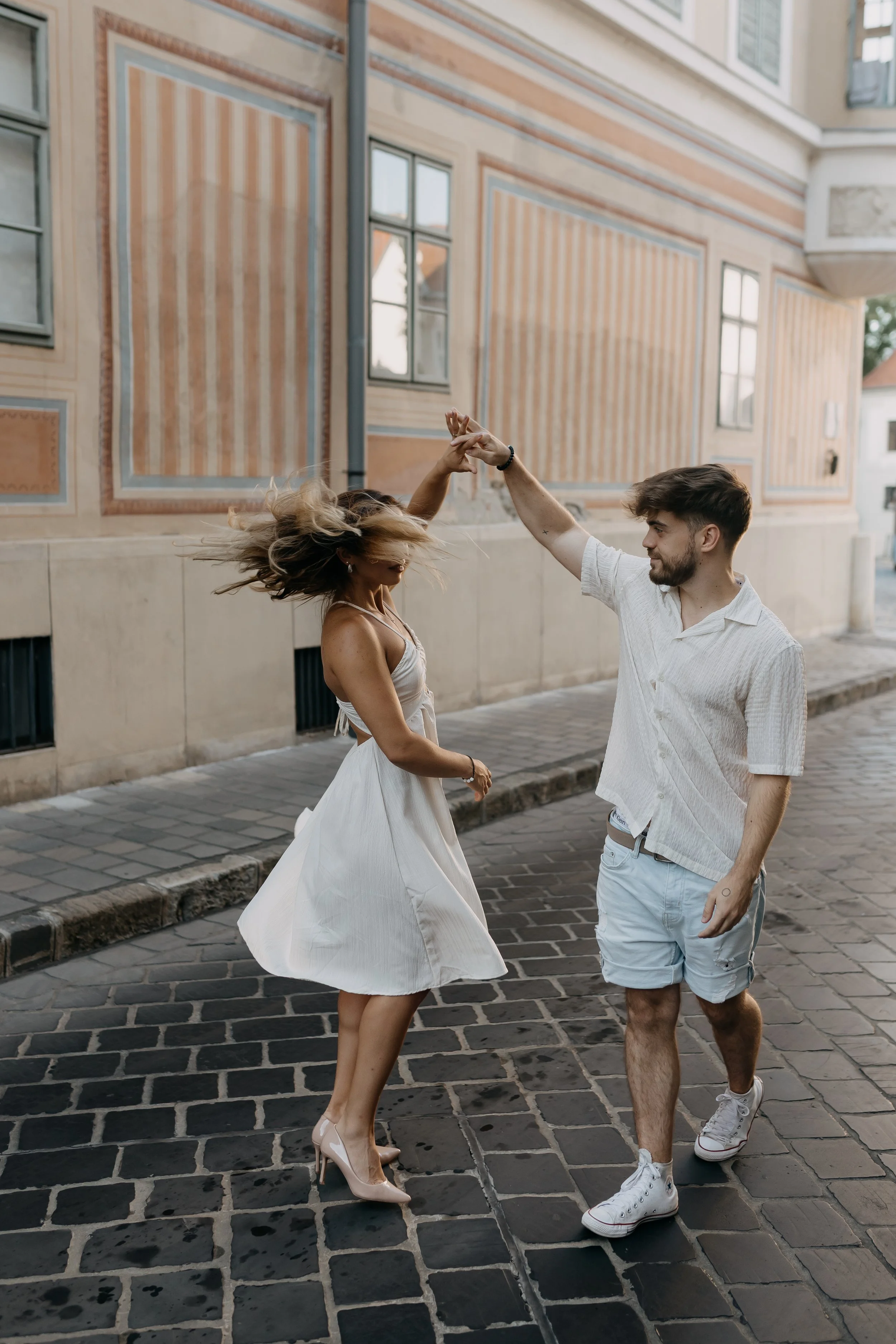 A young man and woman are dancing and holding hands on a cobblestone street in front of a historic building with decorated walls and windows.