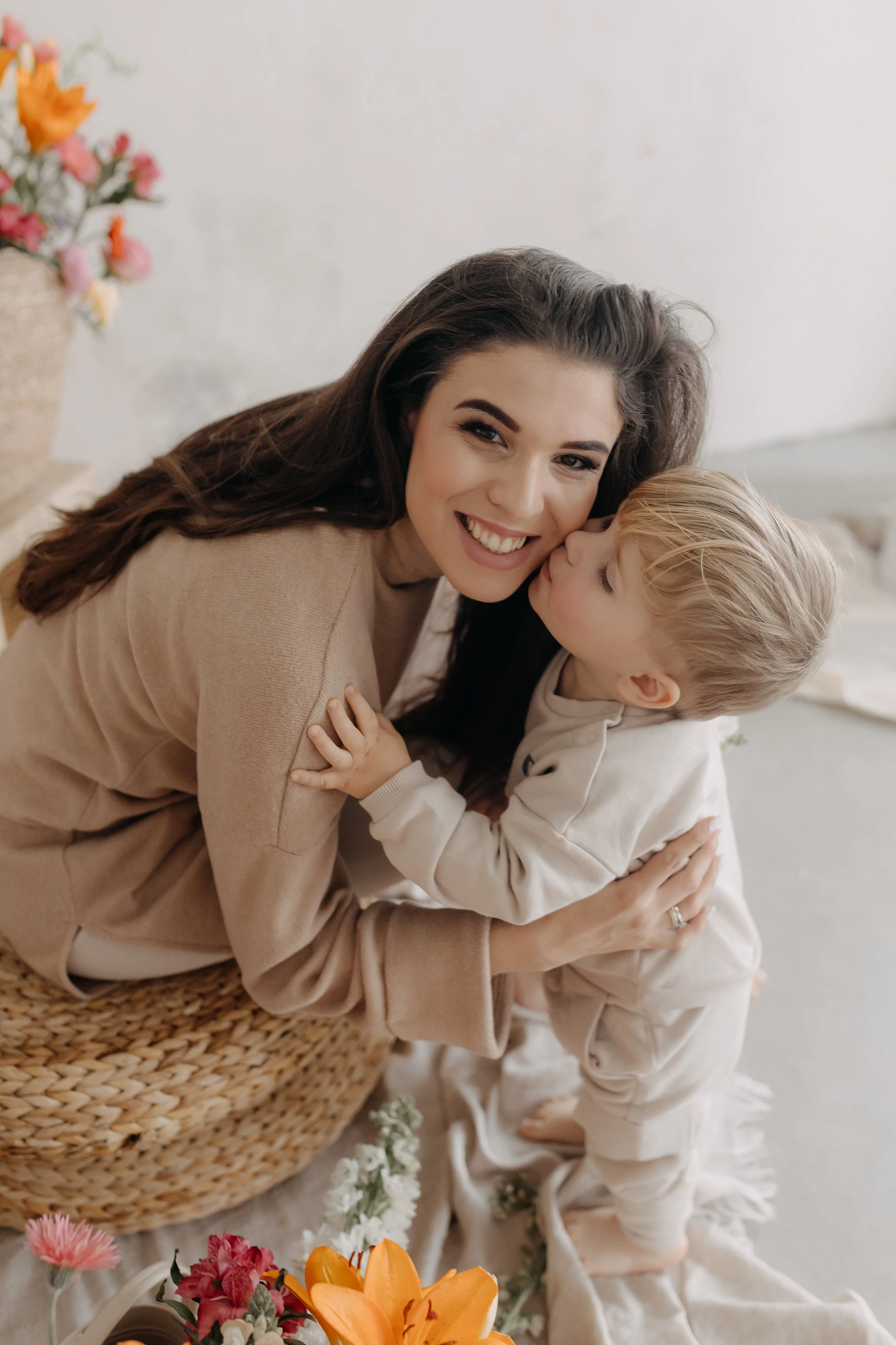 A woman with long dark hair smiling as a young child kisses her cheek, surrounded by flowers and a woven basket.