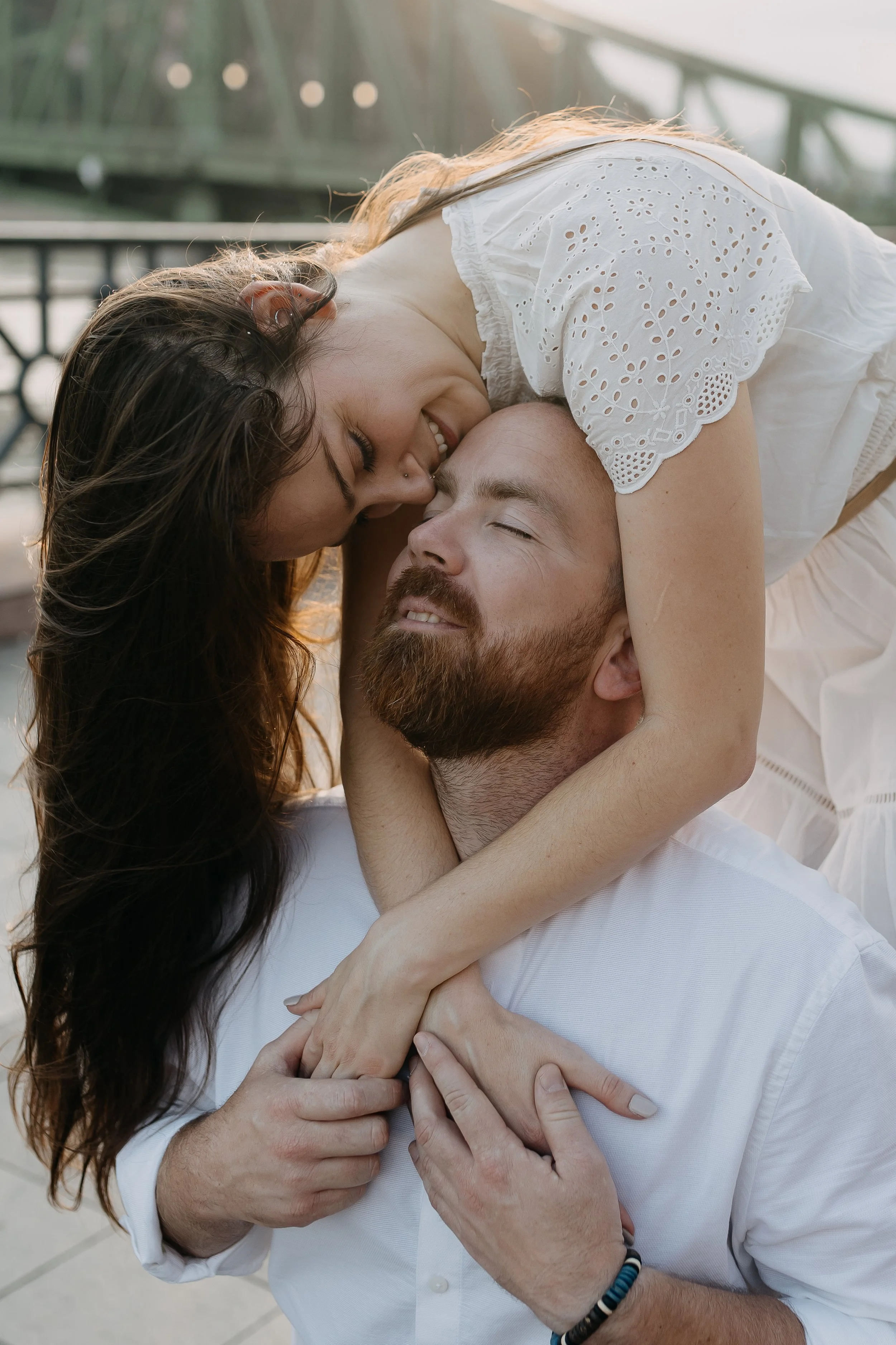 A woman with long dark hair and a man with a beard share a joyful moment, with the woman leaning over the man's head and both embracing while smiling.