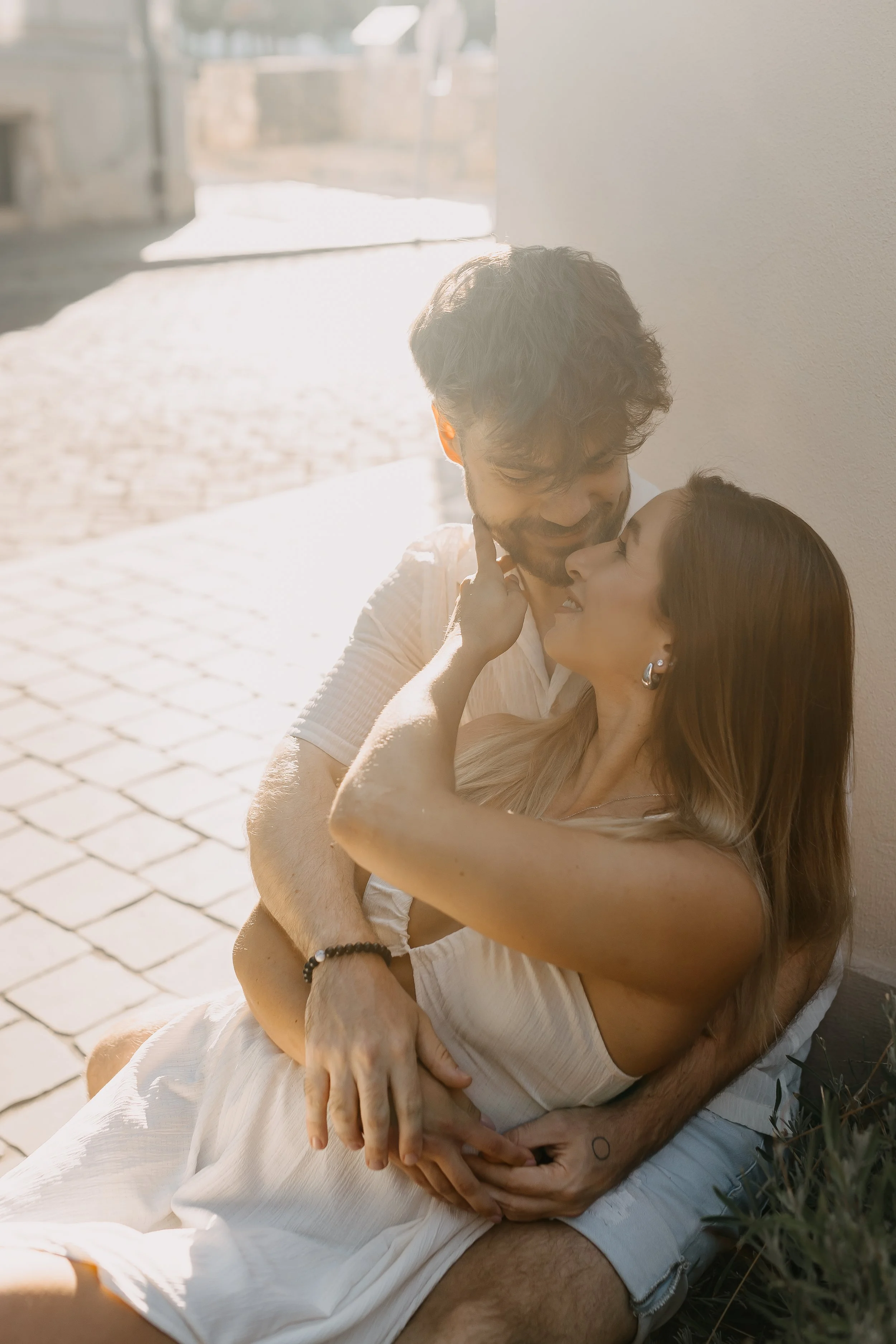 A couple sitting close together on a bench, smiling and looking at each other affectionately in an outdoor setting with sunlight.
