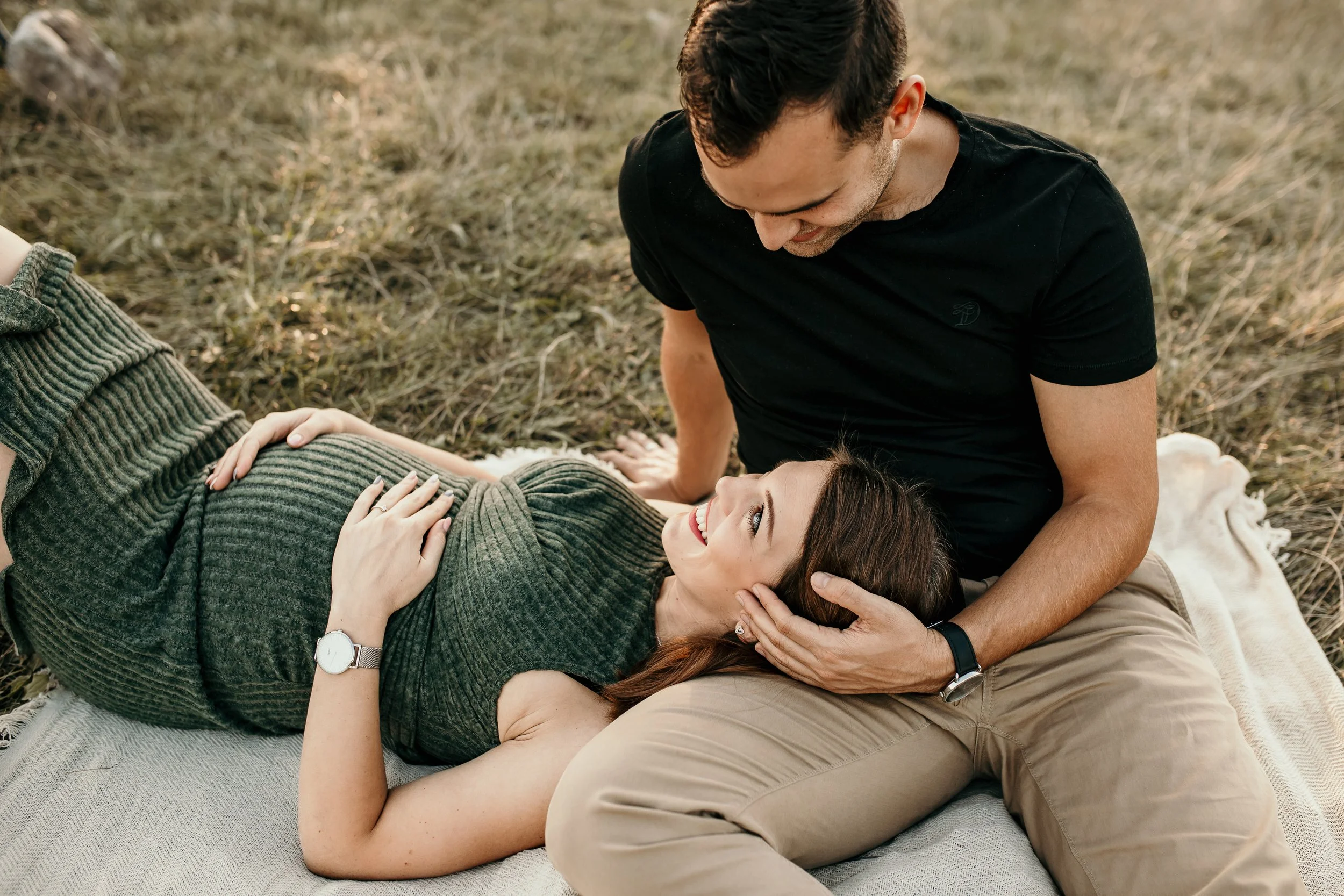 A couple relaxing outdoors on a blanket in a grassy field. The woman is lying on her back, smiling, with her arm resting on her stomach, while the man is sitting beside her, leaning over and looking at her with a smile, gently holding her head.