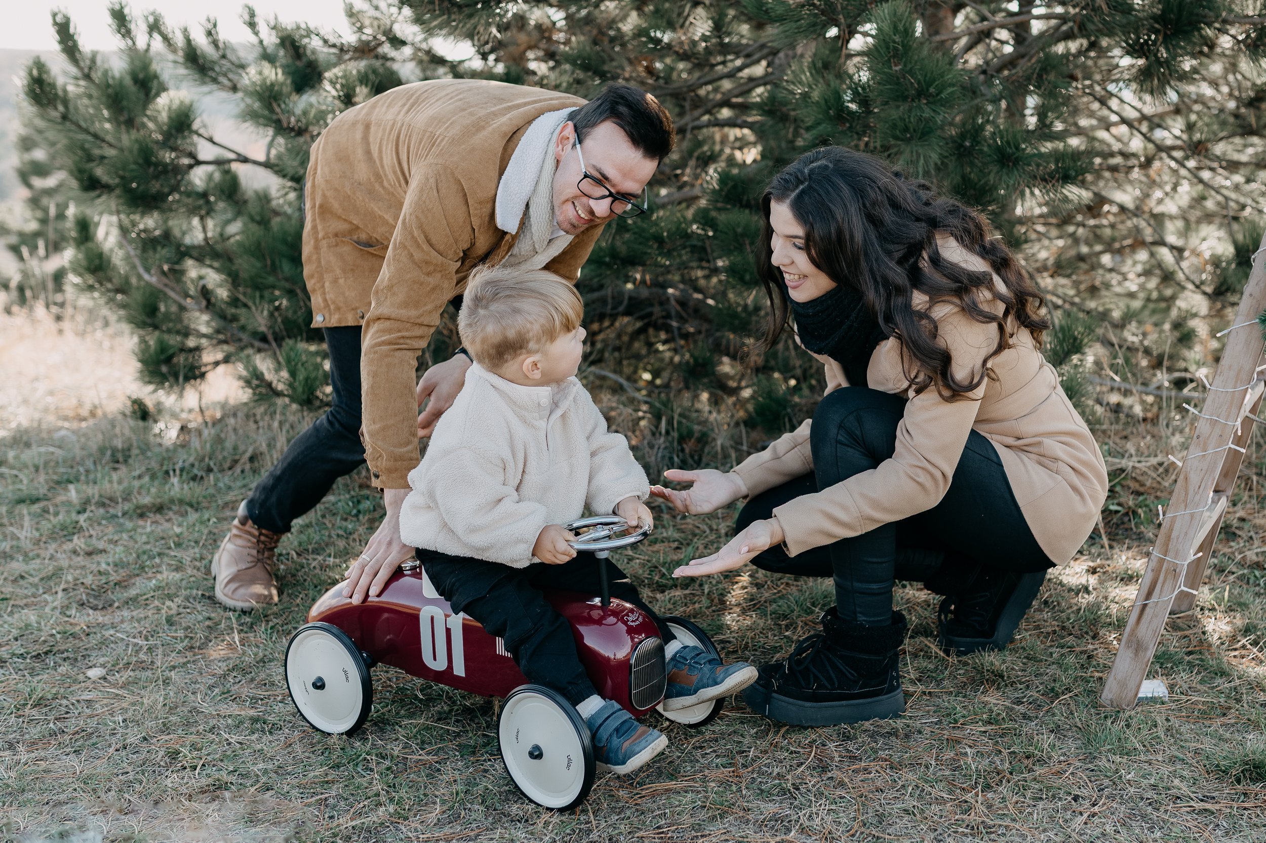 A family of three outdoors with pine trees, a man, woman, and young boy, playing together. The boy is sitting on a small red ride-on toy car while the woman kneels next to him, holding his hand. The man is bending over, smiling at the children. The family appears happy and engaged in a playful moment.