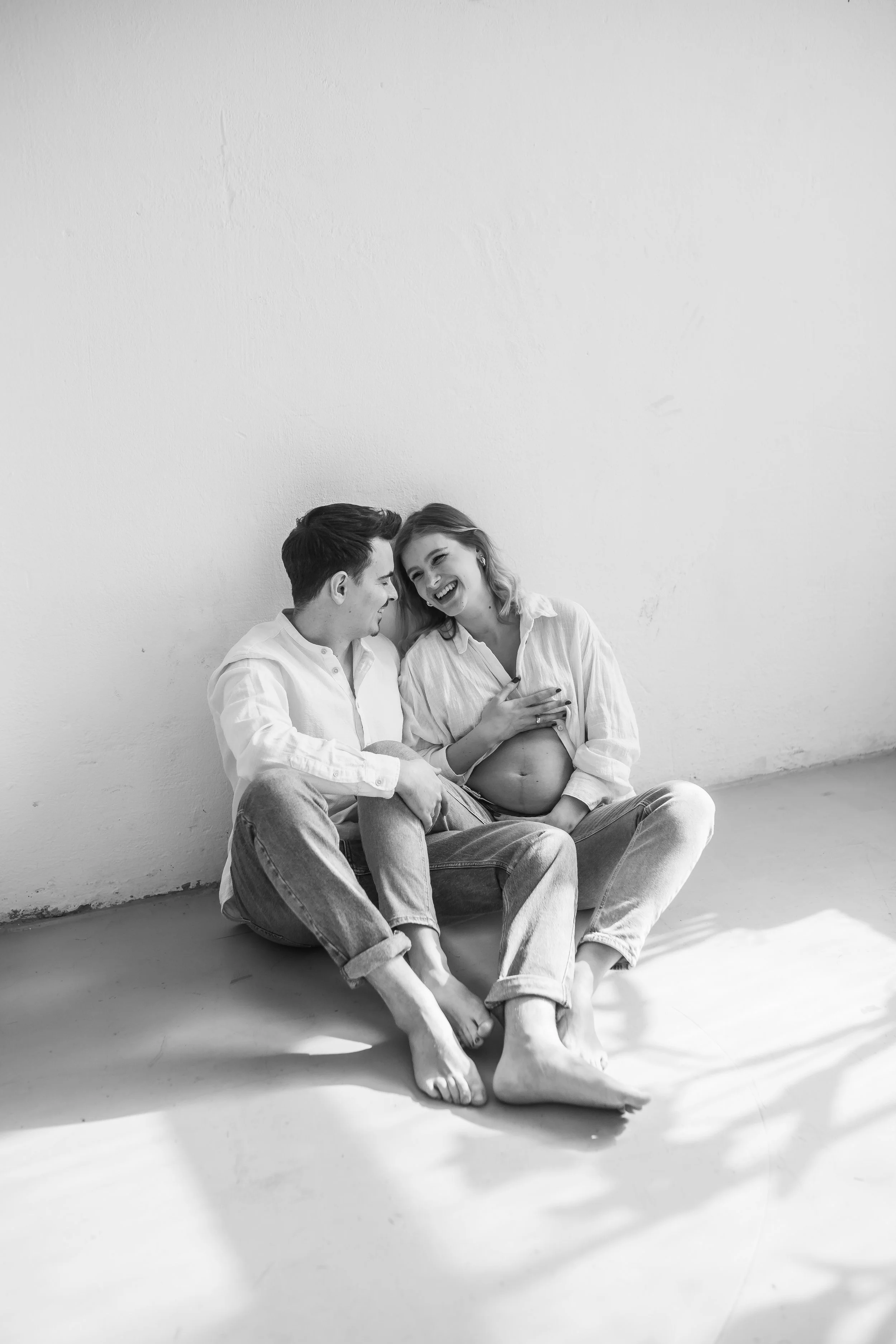 Black and white photo of a smiling man and woman sitting on the floor against a plain wall, with the woman showing her pregnant belly and the man holding her hand.