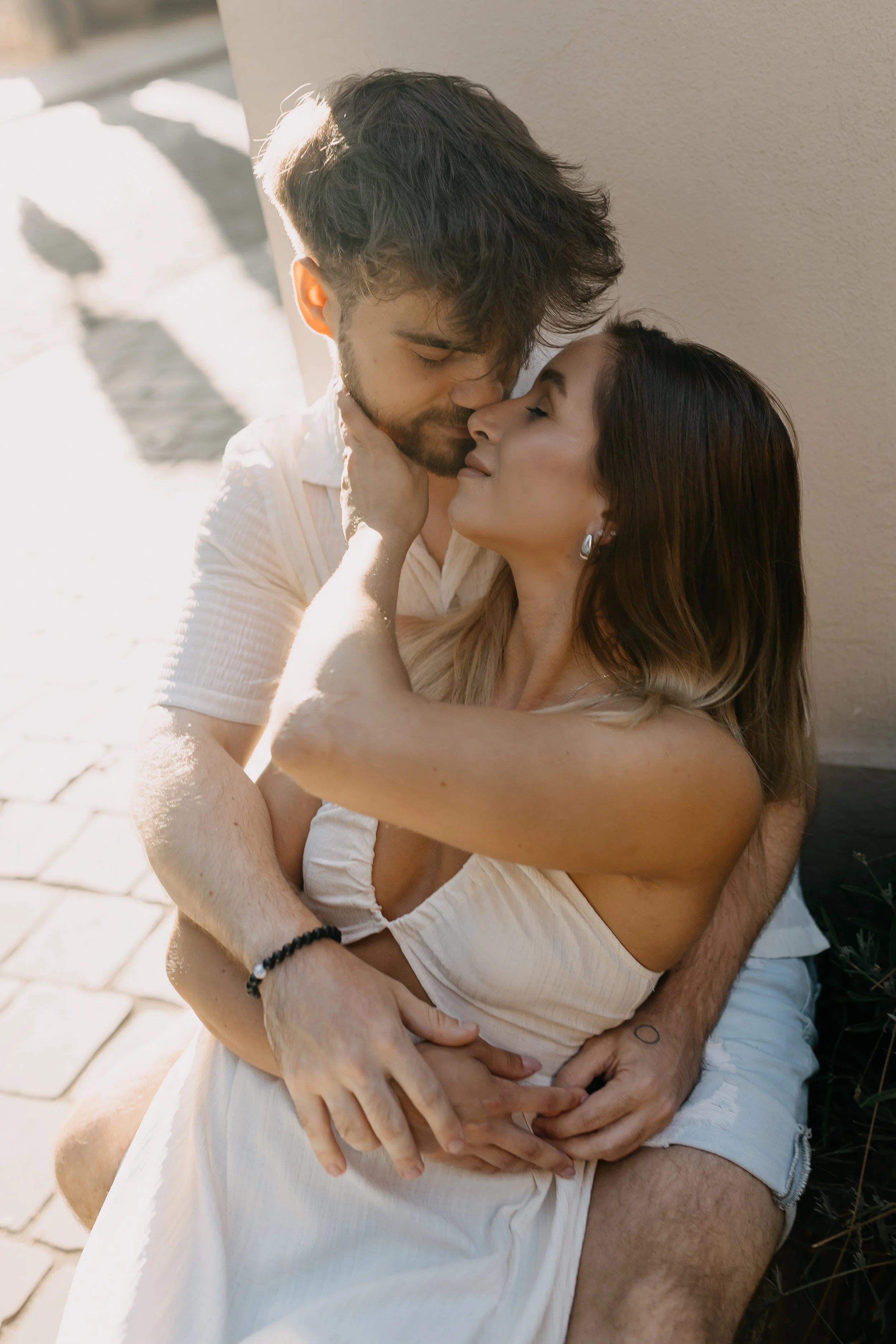 A couple sharing an intimate moment outdoors, with the man holding the woman in his arms as they touch foreheads and close their eyes, basking in sunlight.