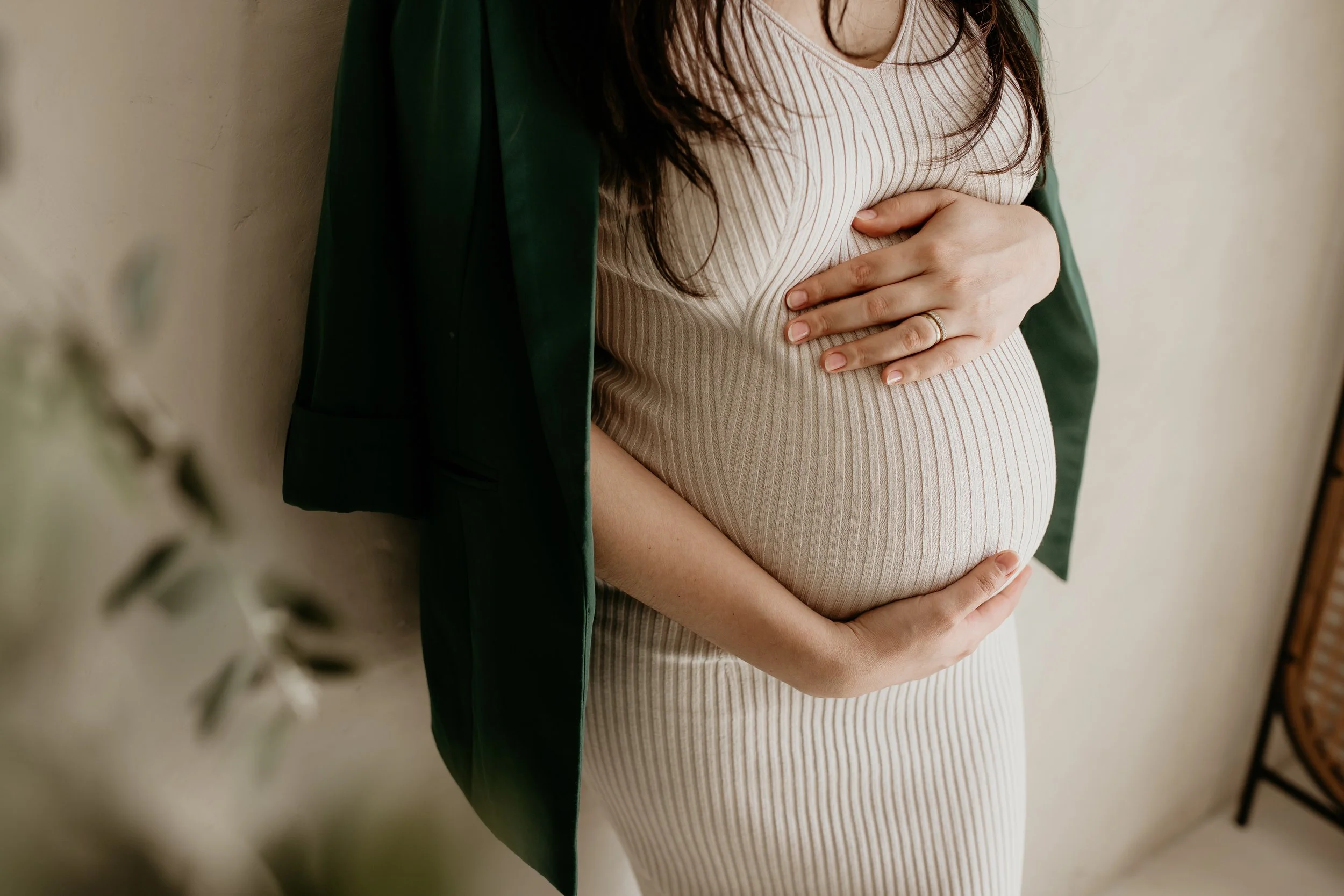 A pregnant woman with dark hair wearing a striped beige dress and a green blazer, standing indoors, cradling her belly with her hands.