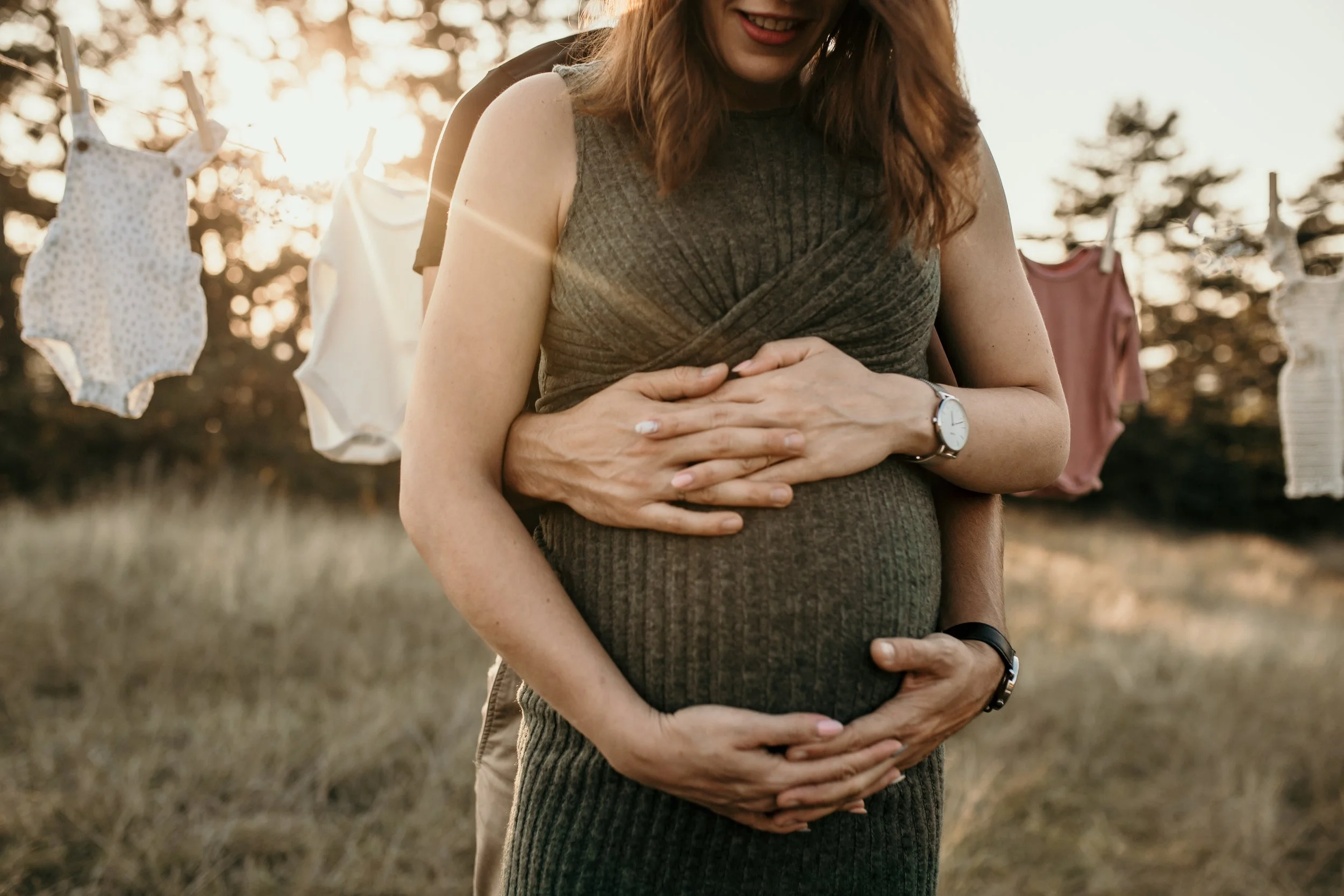 A pregnant woman and a man are standing outdoors in a field, with the woman in front and the man behind her, both holding her belly. The scene is backlit by the setting sun, creating a warm glow. There are clothes hanging on a line in the background.