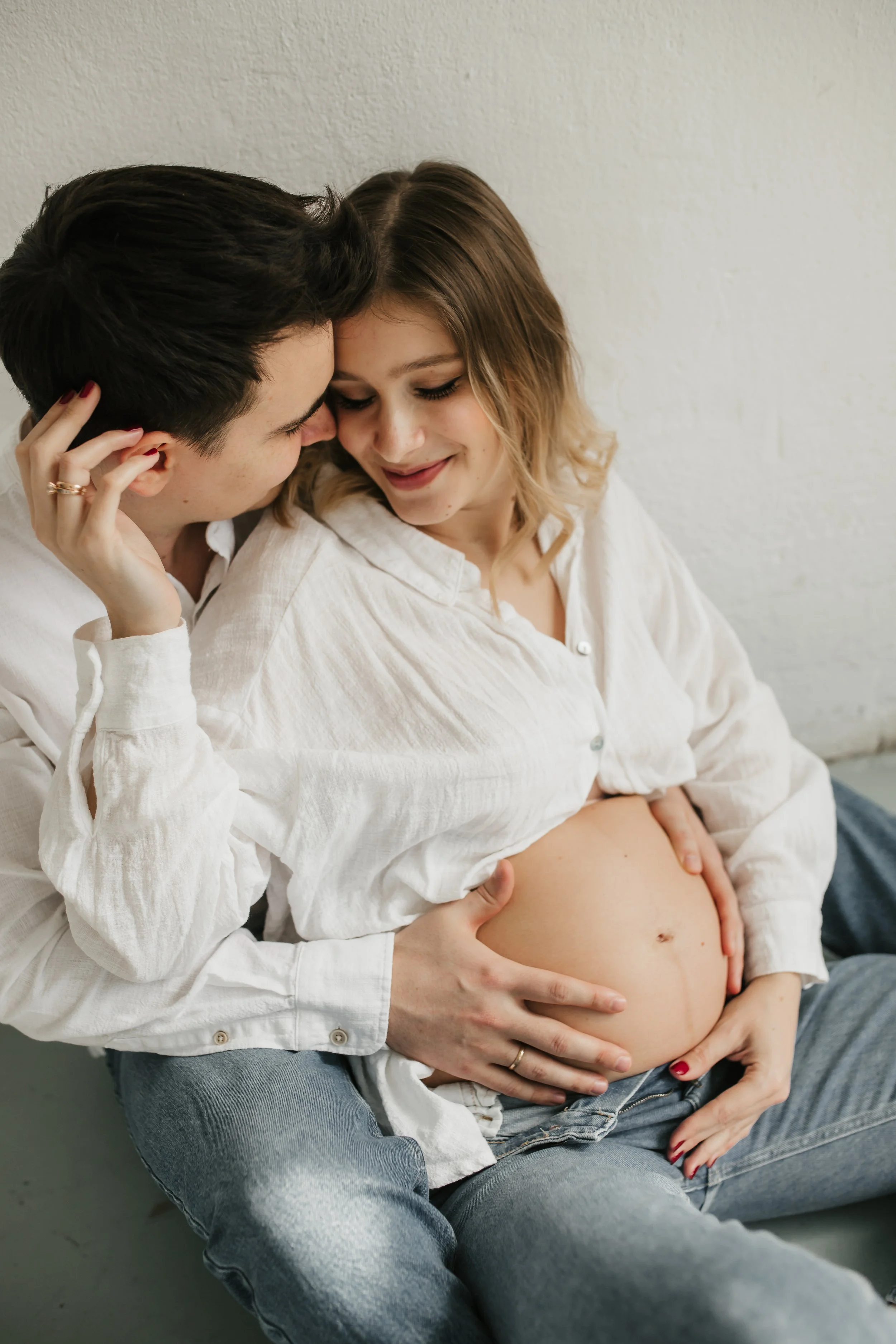 A pregnant woman sitting on a person's lap, with a man holding her belly and gently touching her arm, both smiling softly.