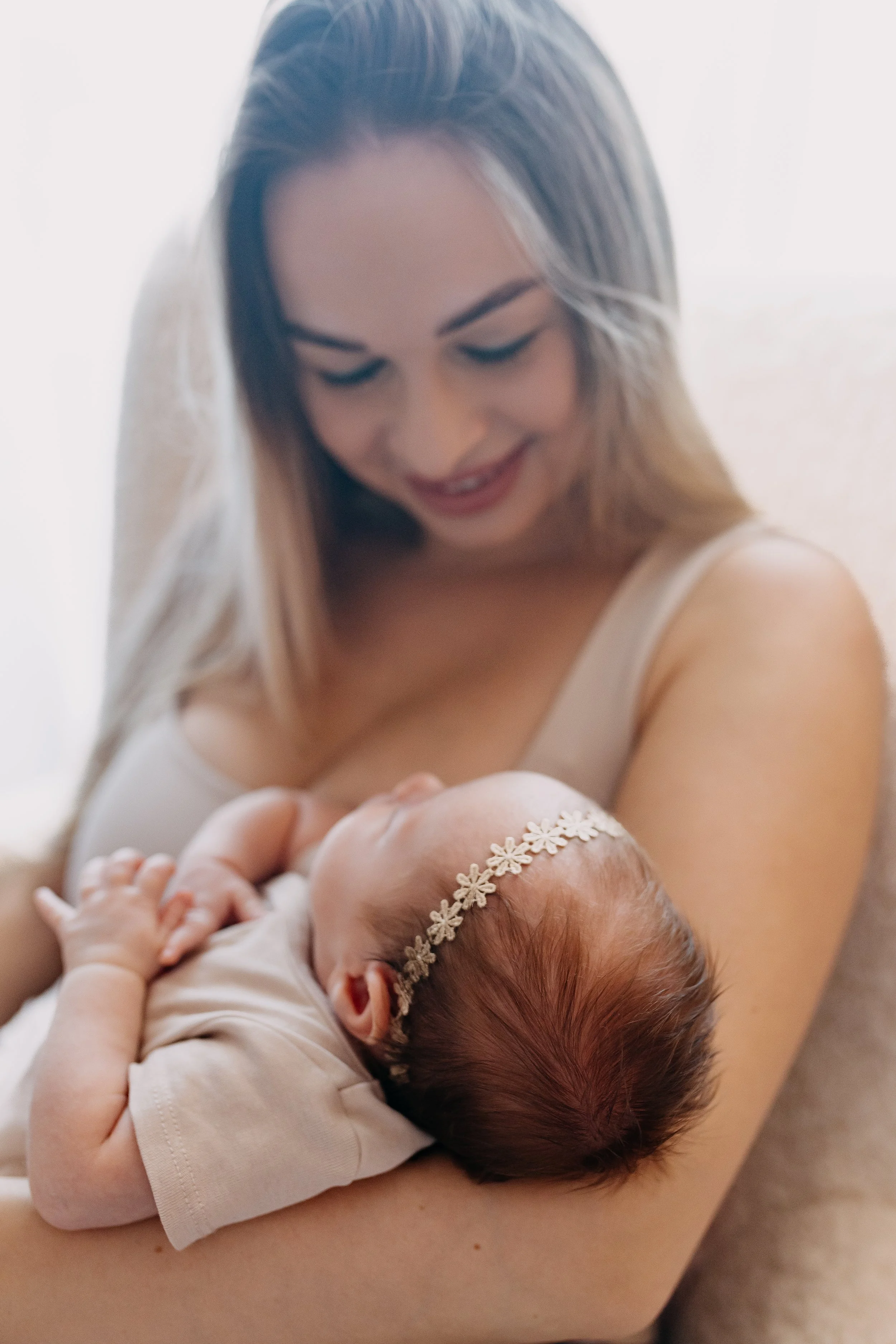 A woman is smiling and looking down at a sleeping baby she is holding. The baby is wearing a floral headband and a white outfit.