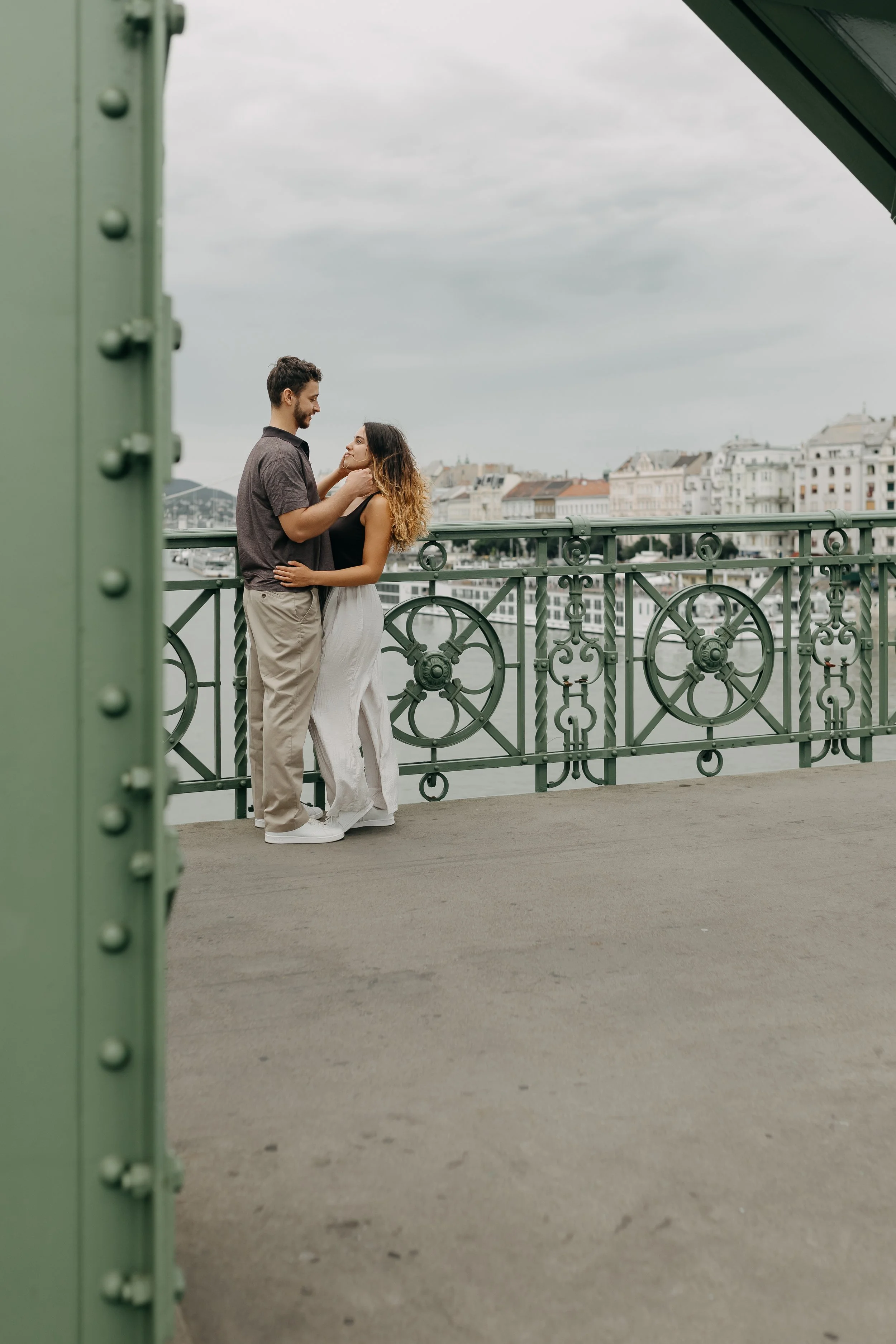 A young couple with light skin and average build sharing a romantic moment on a bridge with a cityscape in the background. The man, wearing a dark short-sleeve shirt and beige pants, gently holds the woman's face and waist. The woman, dressed in a bl