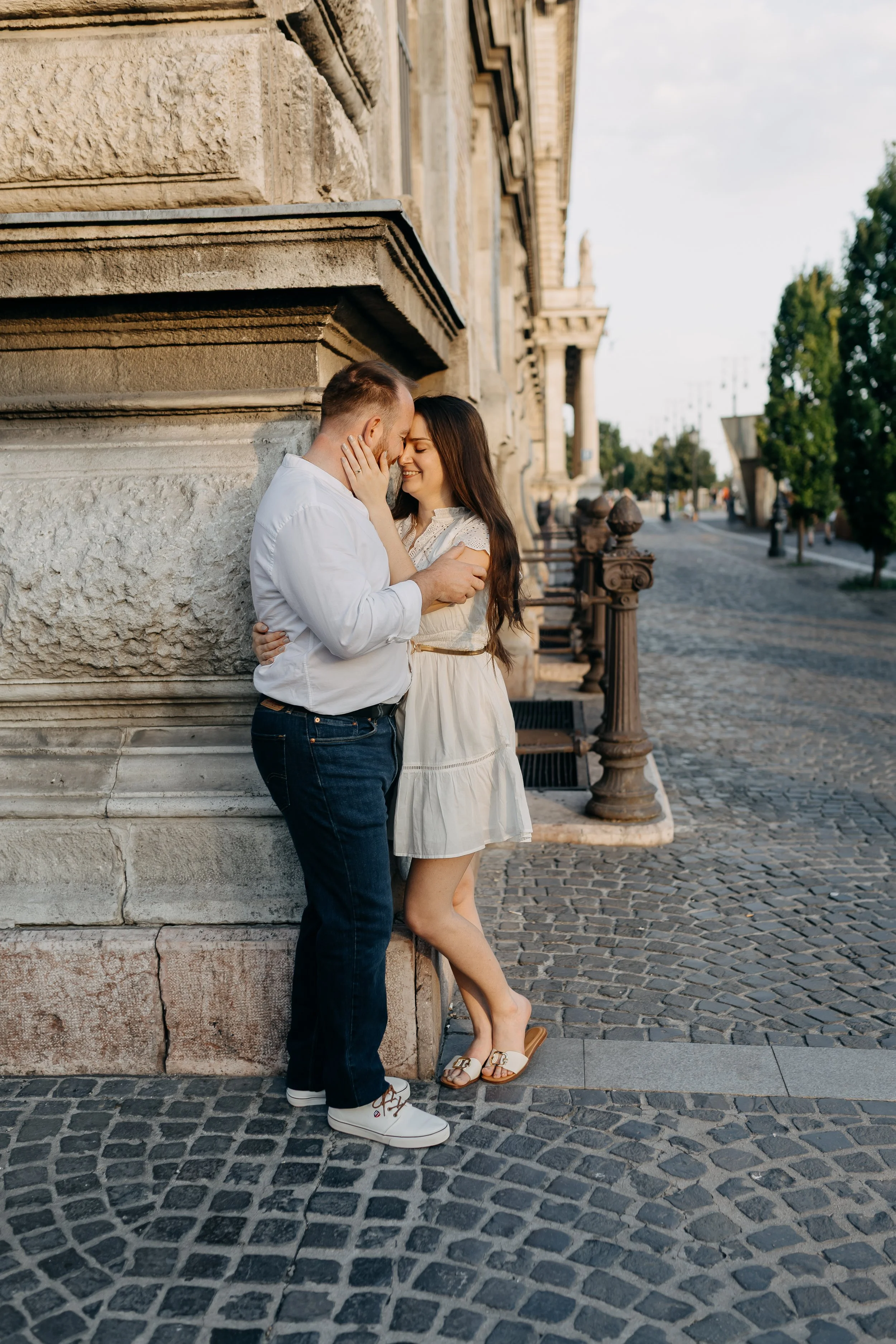 A young couple sharing an affectionate moment on a cobblestone street beside a stone building with architectural details. The woman, wearing a white dress and sandals, is leaning against the wall with her feet slightly lifted. The man, dressed in a w