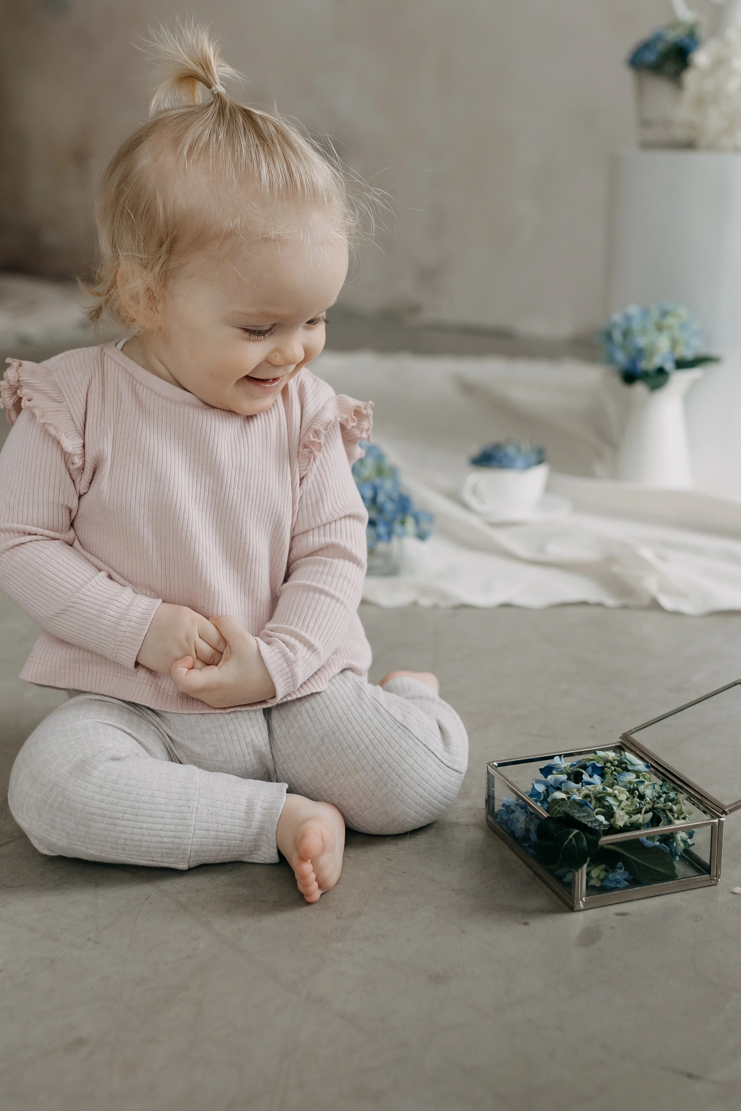 A young girl with blonde hair, seated cross-legged on a beige carpet, smiling and looking downward. She is wearing a light pink, ruffled long-sleeve top and beige pants. In front of her is a glass box filled with blue and green artificial flowers. Th