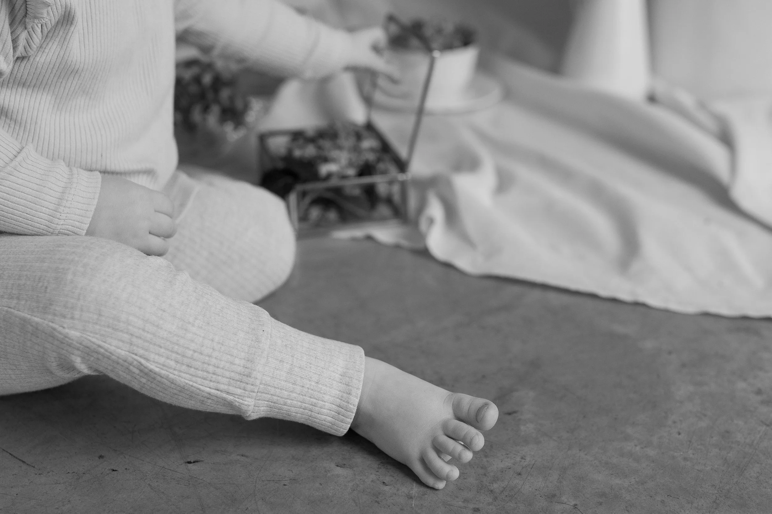 A close-up view of a child's feet and lower legs as they sit on the floor, with one hand resting on their knee. The child is wearing light-colored, textured pants and a long-sleeved top. In the background, there is a small, open box filled with vario