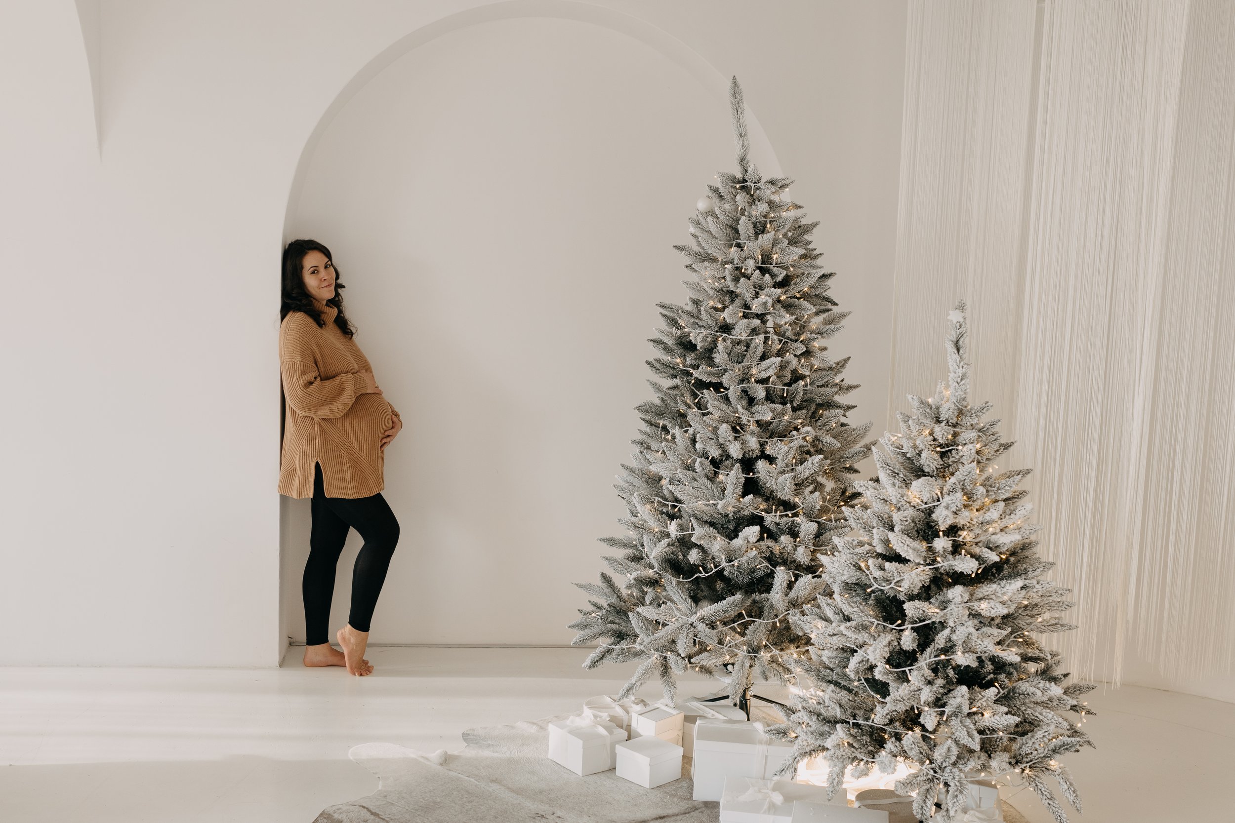 A pregnant woman standing barefoot next to a white Christmas tree with snow-like branches, decorated with warm white lights, in a minimalist room with white walls and wood paneling. Several wrapped presents are placed under the tree.
