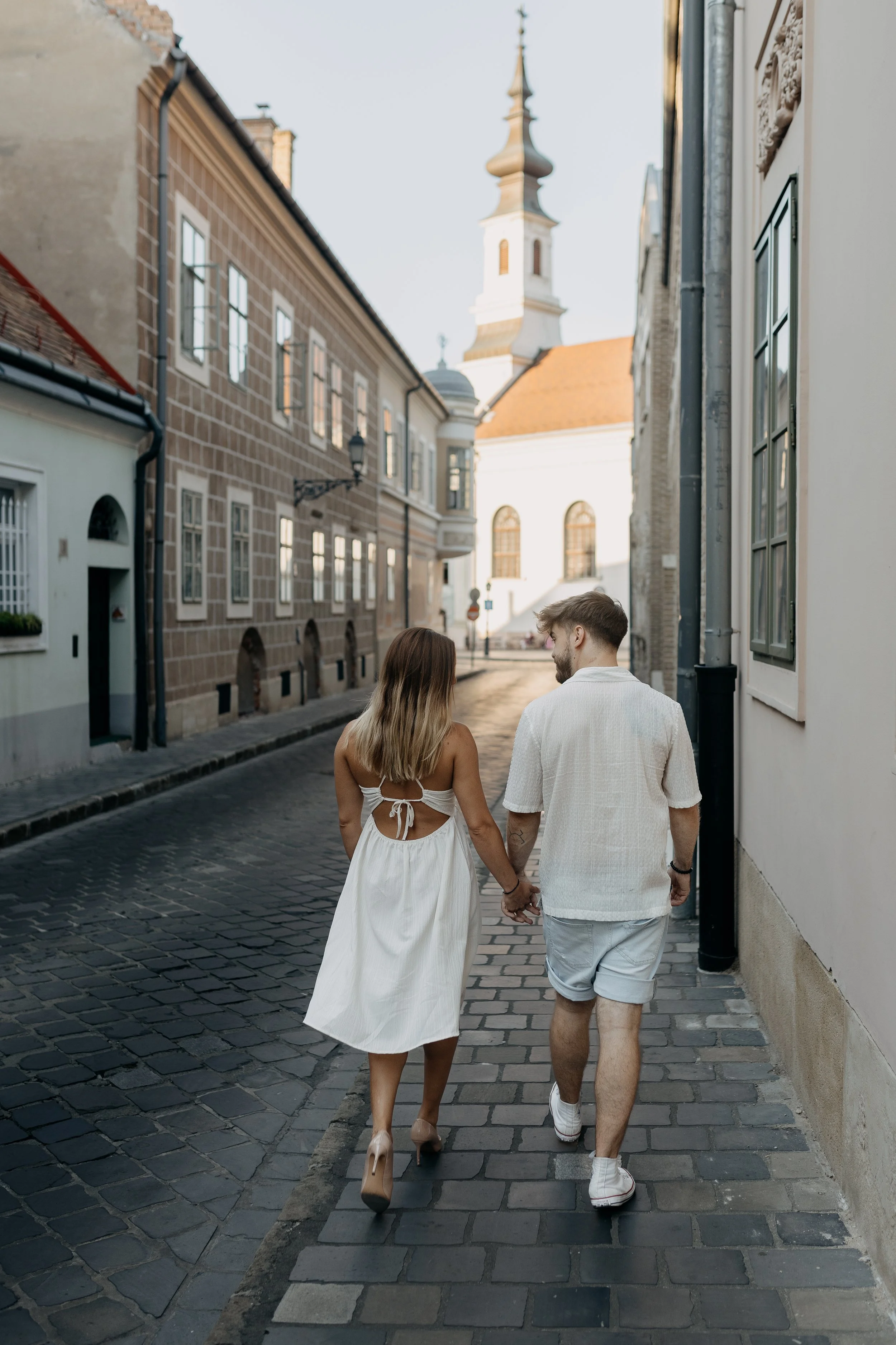 A couple holding hands and walking down a cobblestone street in a European town, with historic buildings in the background.
