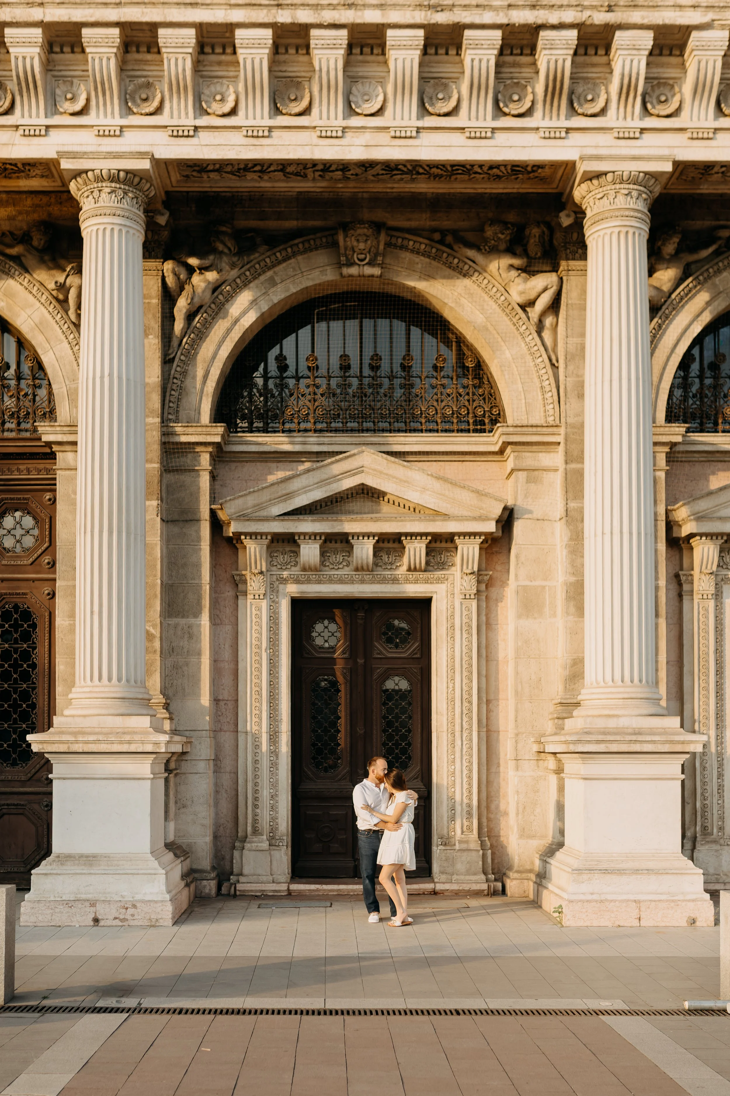 A couple shares a kiss in front of a historic stone building with ornate architectural details and columns.