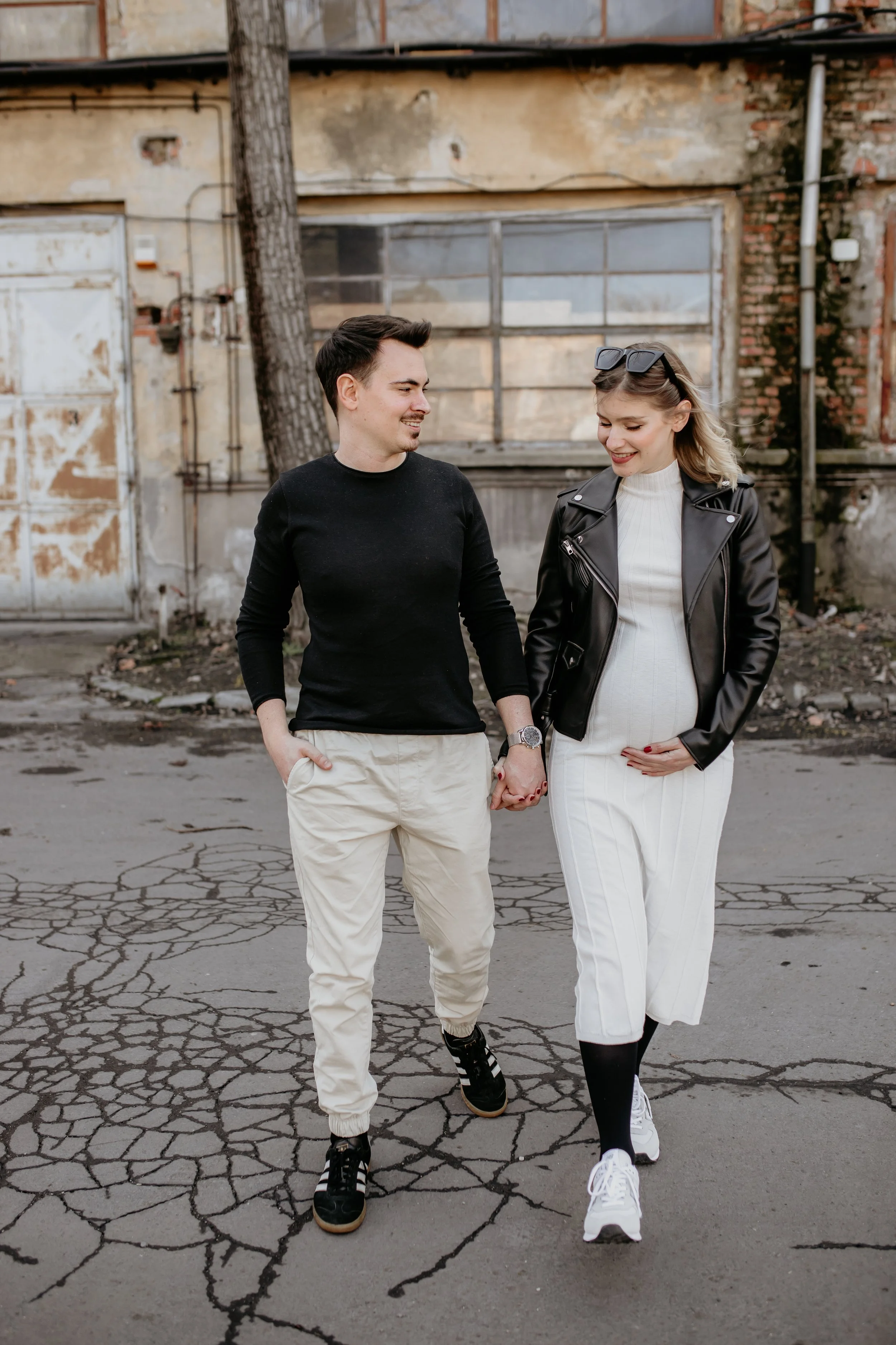 a man and woman walking together outside, holding hands, in front of an old brick and concrete wall