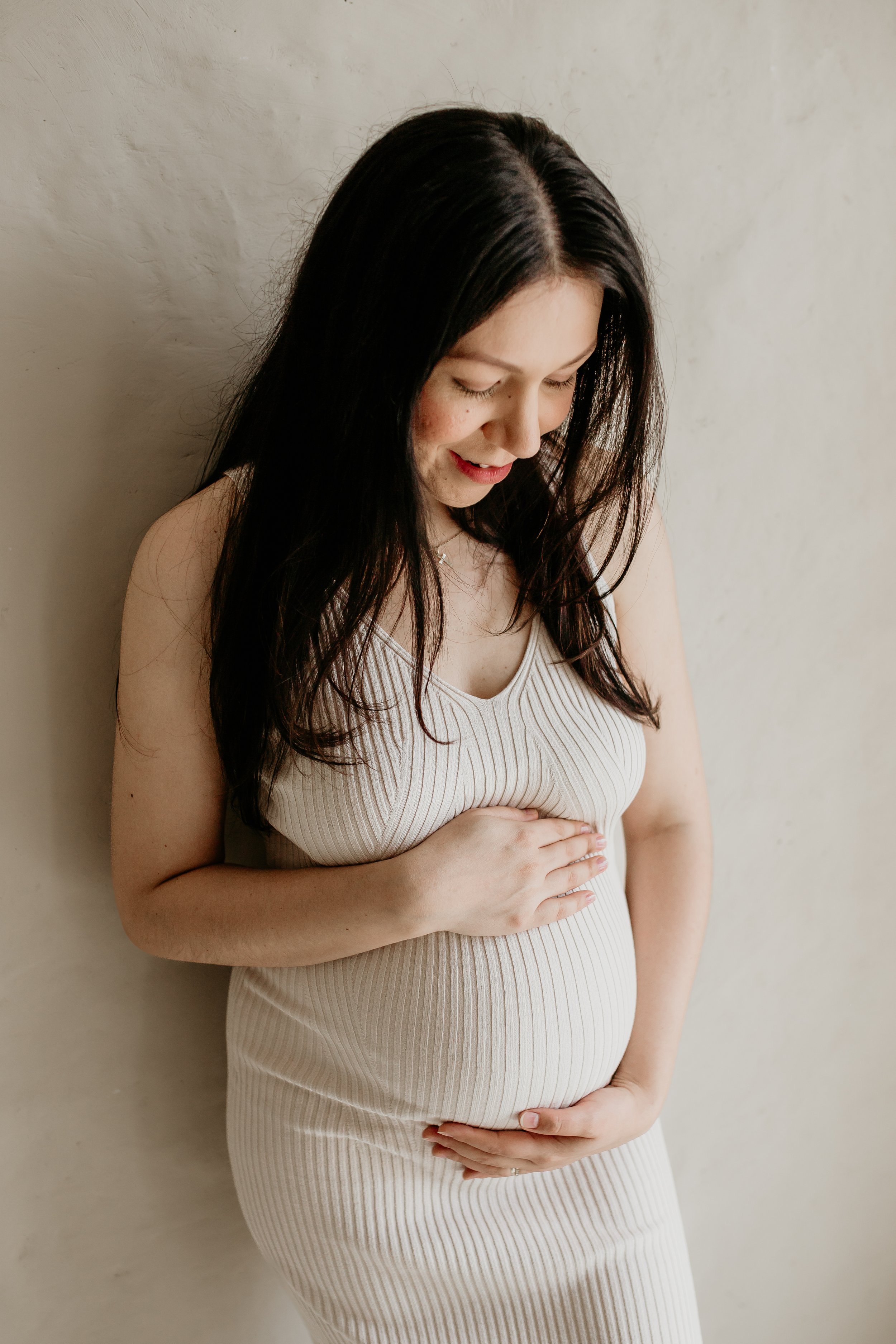 A pregnant woman with long dark hair is standing against a plain wall, smiling and gently holding her baby bump with both hands, wearing a beige sleeveless ribbed dress.