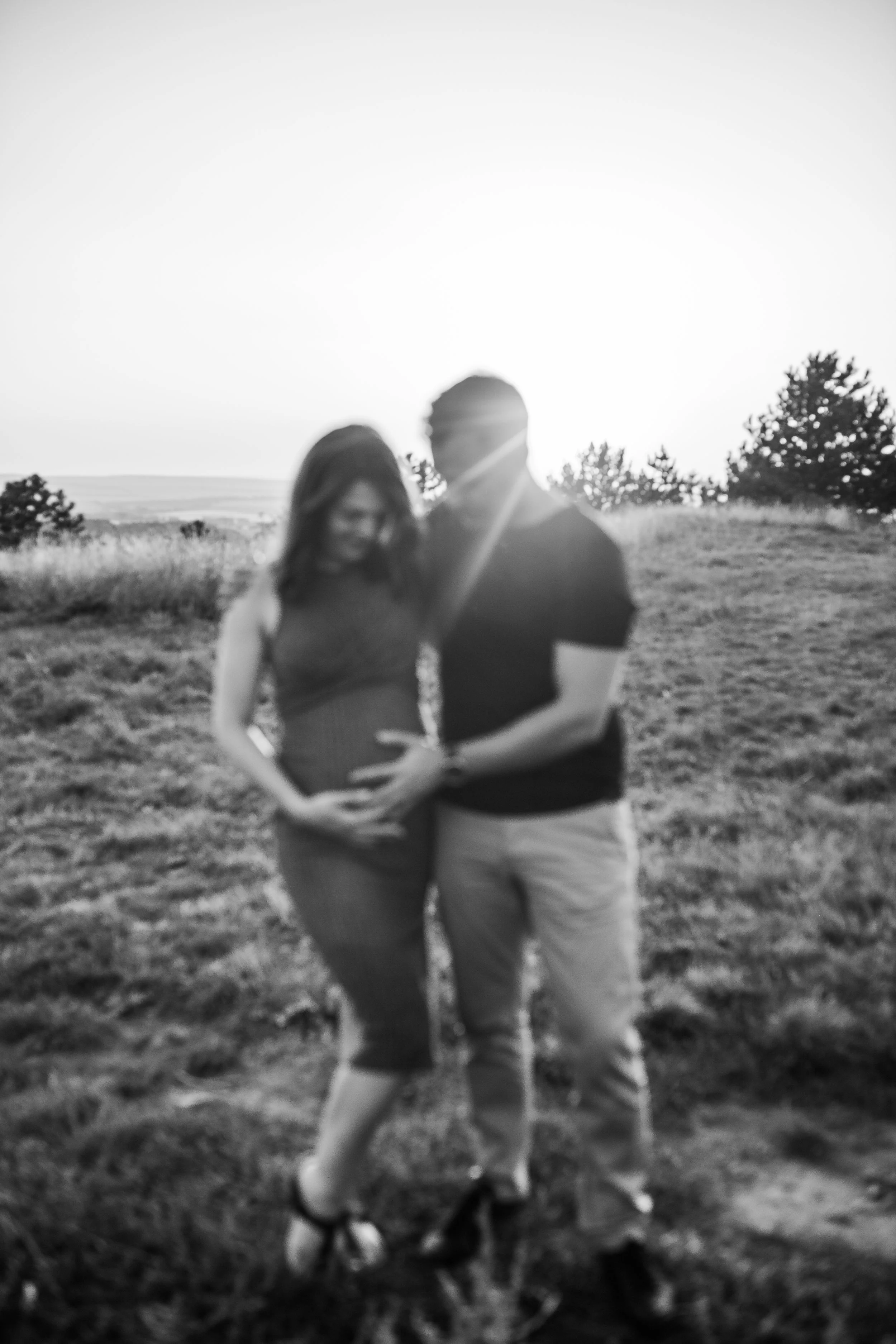 A black-and-white photo of a pregnant woman smiling and a man with their heads close together, standing outdoors in a natural setting with trees and open sky in the background. They are holding hands around her stomach.