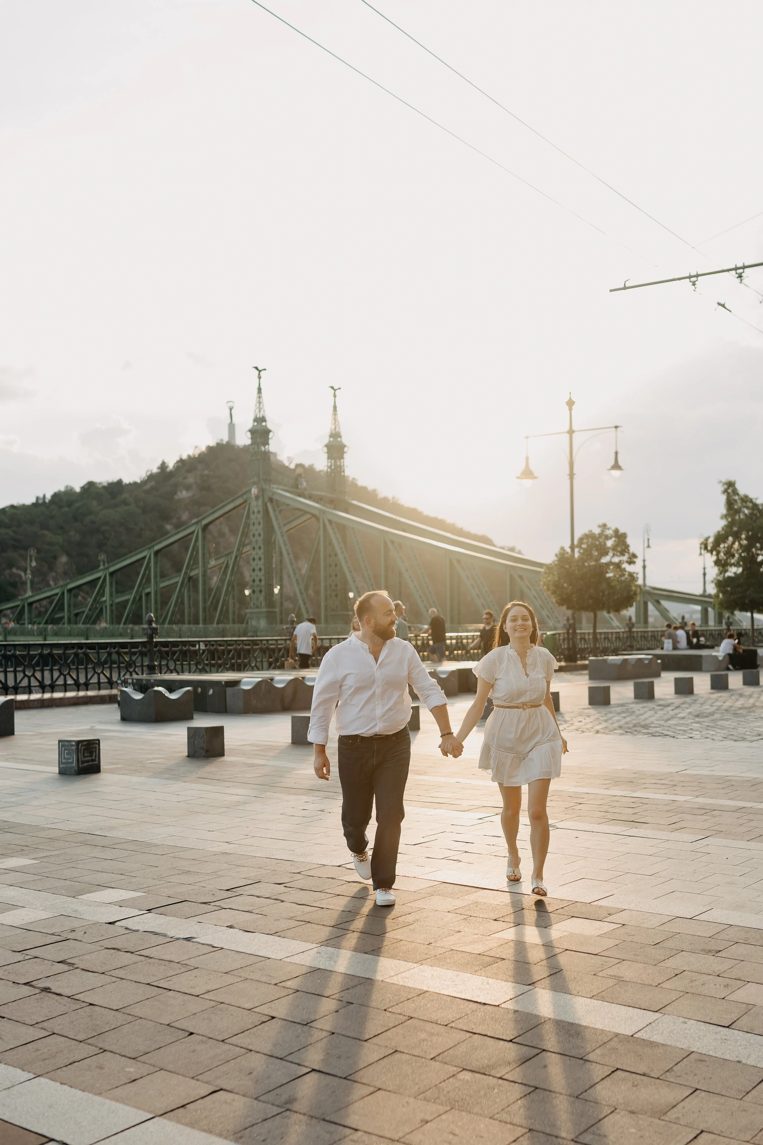 A couple holding hands and walking on a city street at sunset, with a bridge, streetlights, and a hill with towers in the background.