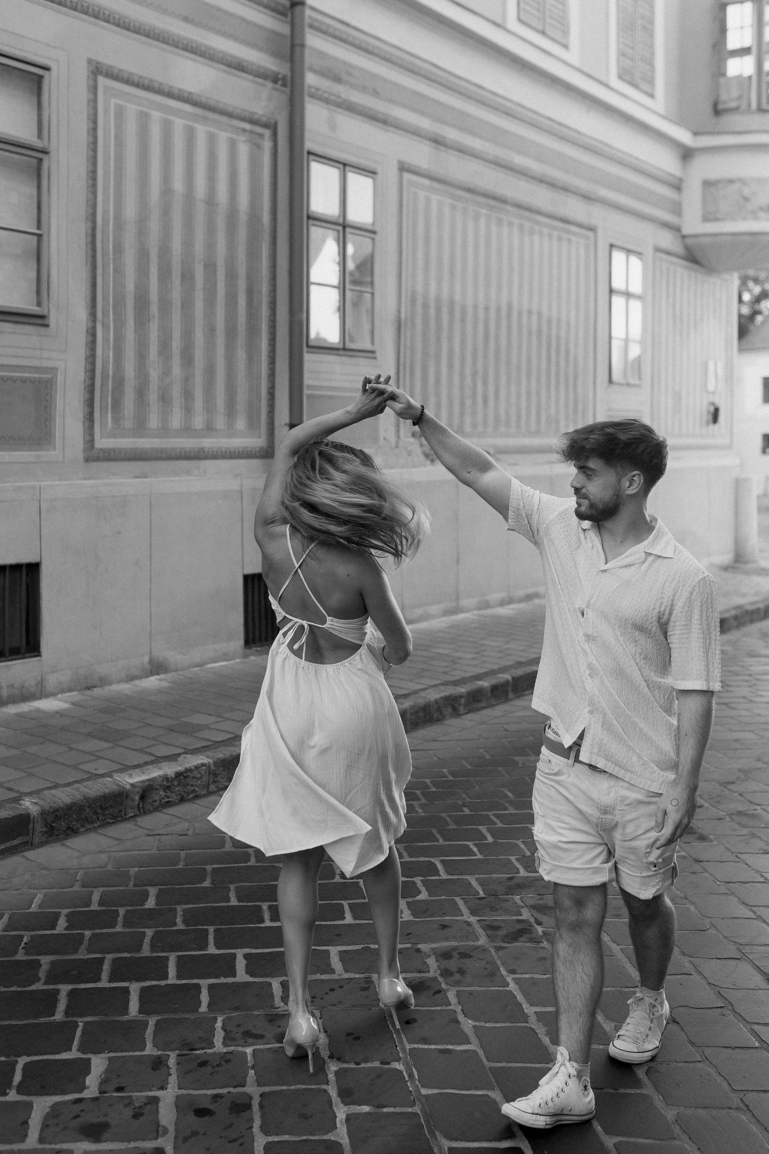A young man and woman dancing on a cobblestone street in front of a building with large windows. The woman is spinning, wearing a light dress, high heels, and has headphones around her neck. The man is holding her hand, wearing a light shirt, shorts,