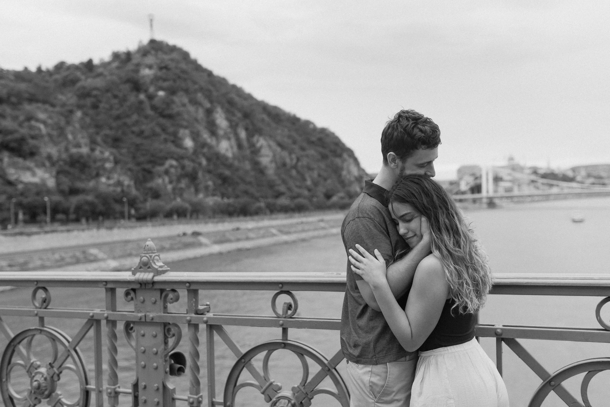 A black and white photo of a man and woman embracing on a bridge with ornate metal railings. The man has short, curly hair and the woman has long, wavy hair. They are smiling and holding each other closely. In the background, there is a hillside with