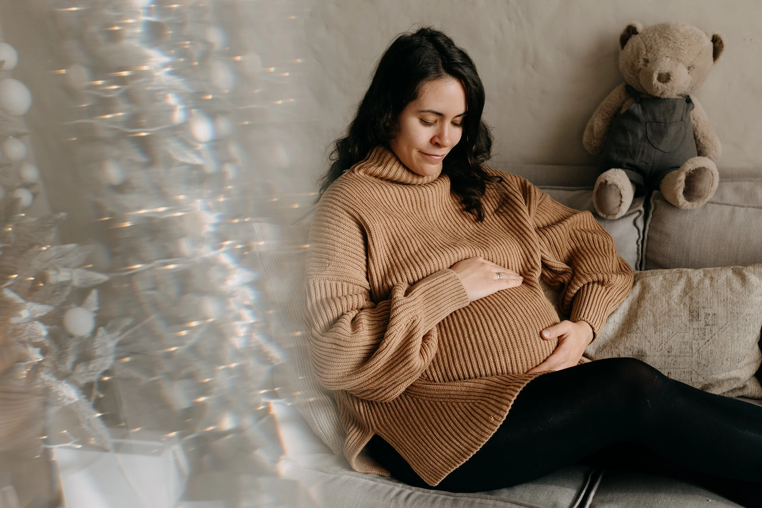 Pregnant woman sitting on a beige sofa, holding her belly, wearing a tan sweater, with a teddy bear sitting beside her, and a reflective light pattern on the left side of the image.