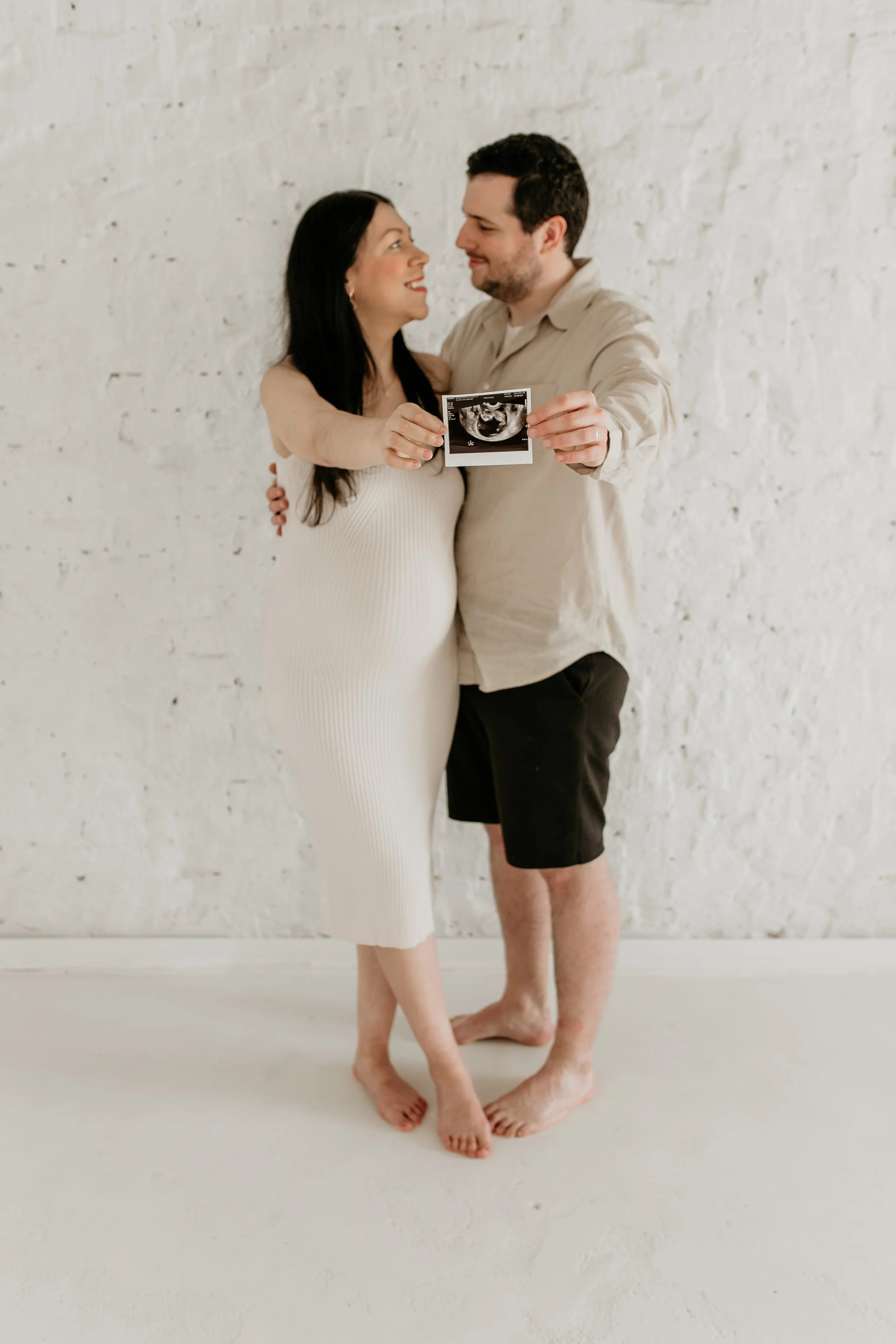 A couple standing close together, holding an ultrasound image, smiling, indoors against a white brick wall.