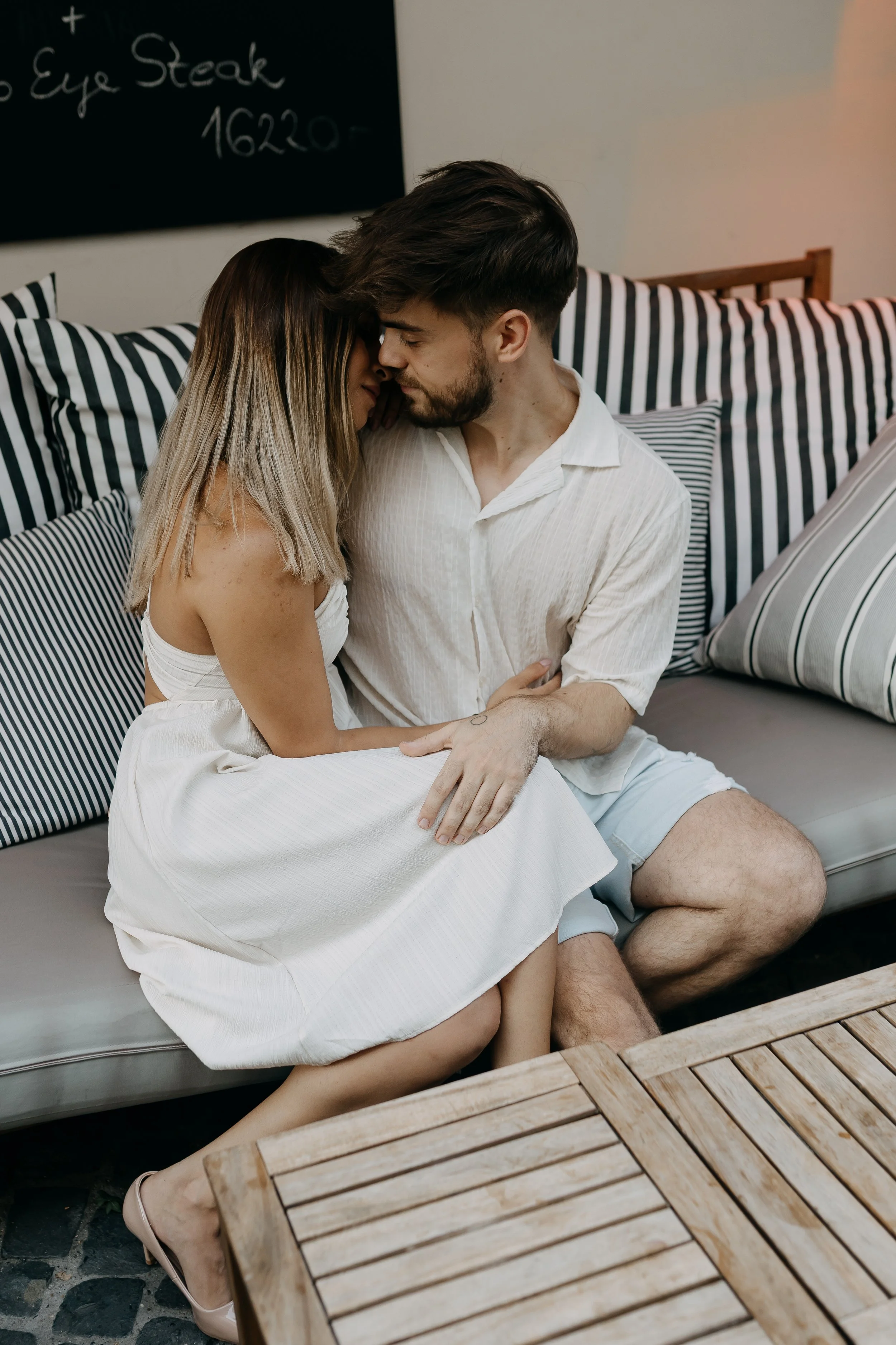 A couple sitting closely on a gray sofa, with their foreheads touching and eyes closed. The woman has long, blonde hair and is wearing a white dress, and the man has short dark hair and a beard, wearing a white shirt and shorts. They are in a cozy in