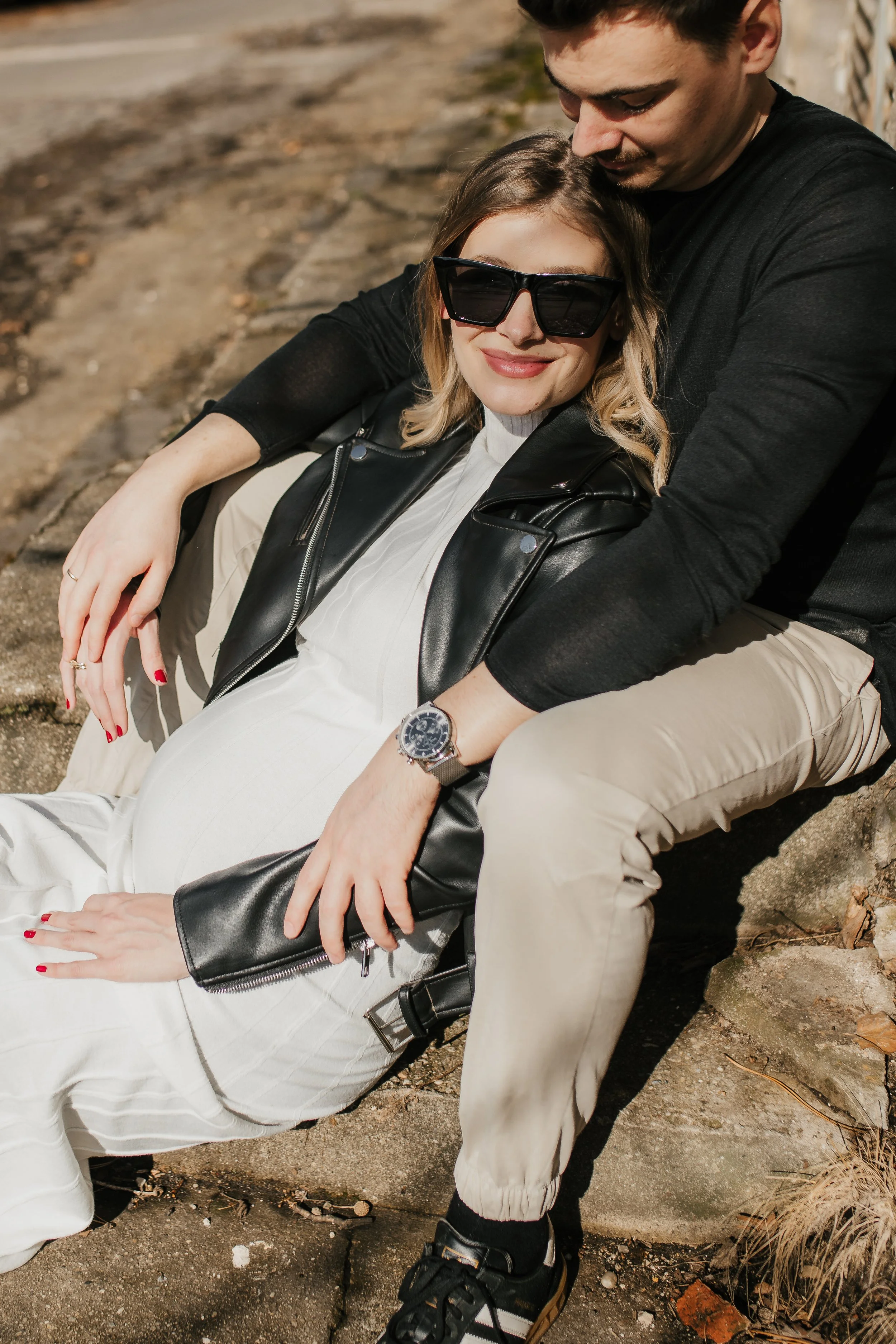A young woman with blonde hair wearing large sunglasses, a white shirt, a black leather jacket, and light-colored pants, smiling and sitting on the ground with a young man behind her. The man has dark hair, is wearing a black shirt and beige pants, a