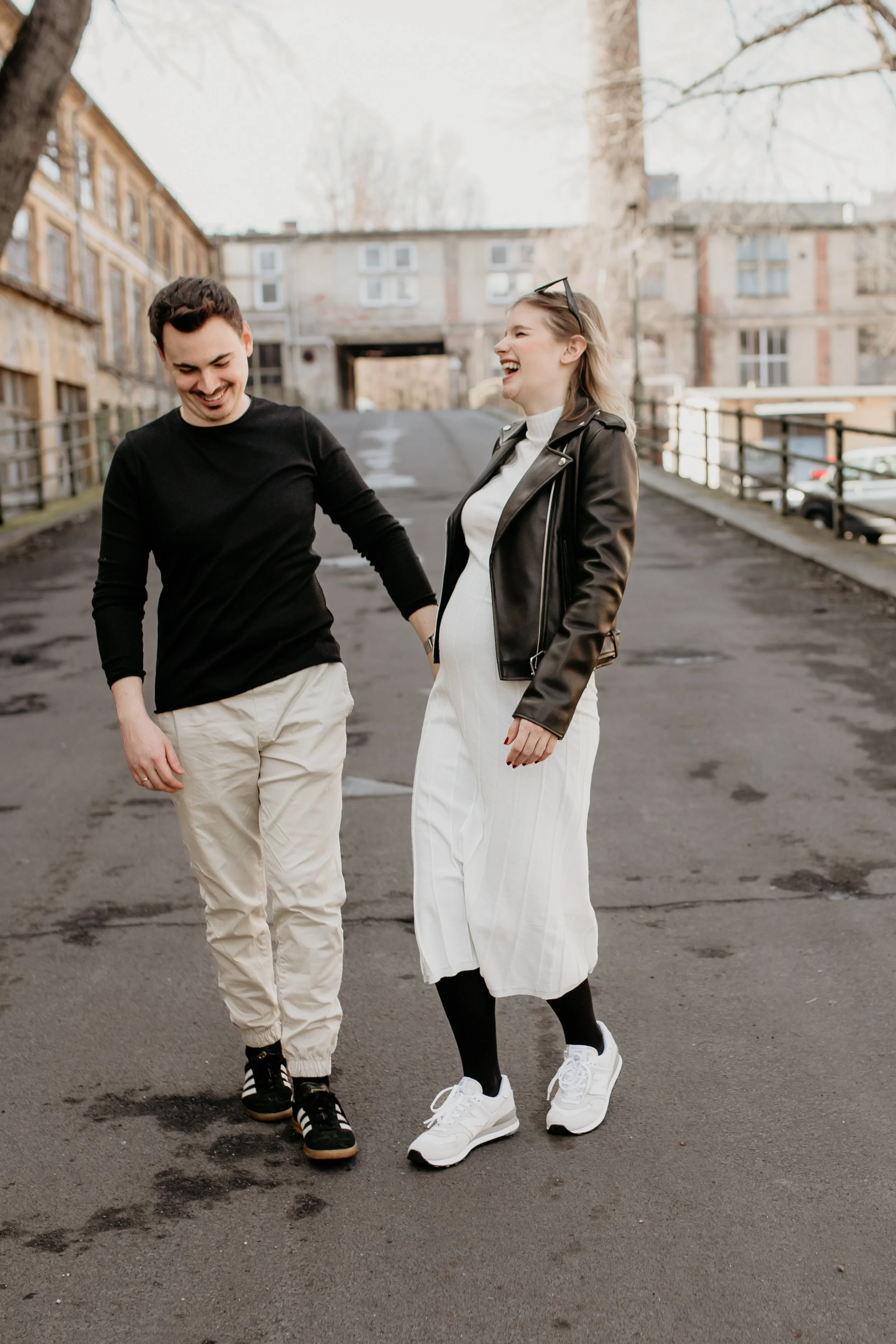 Two young adults, a man and a woman, walking and laughing on a paved street in an urban area, with residential buildings and leafless trees in the background.