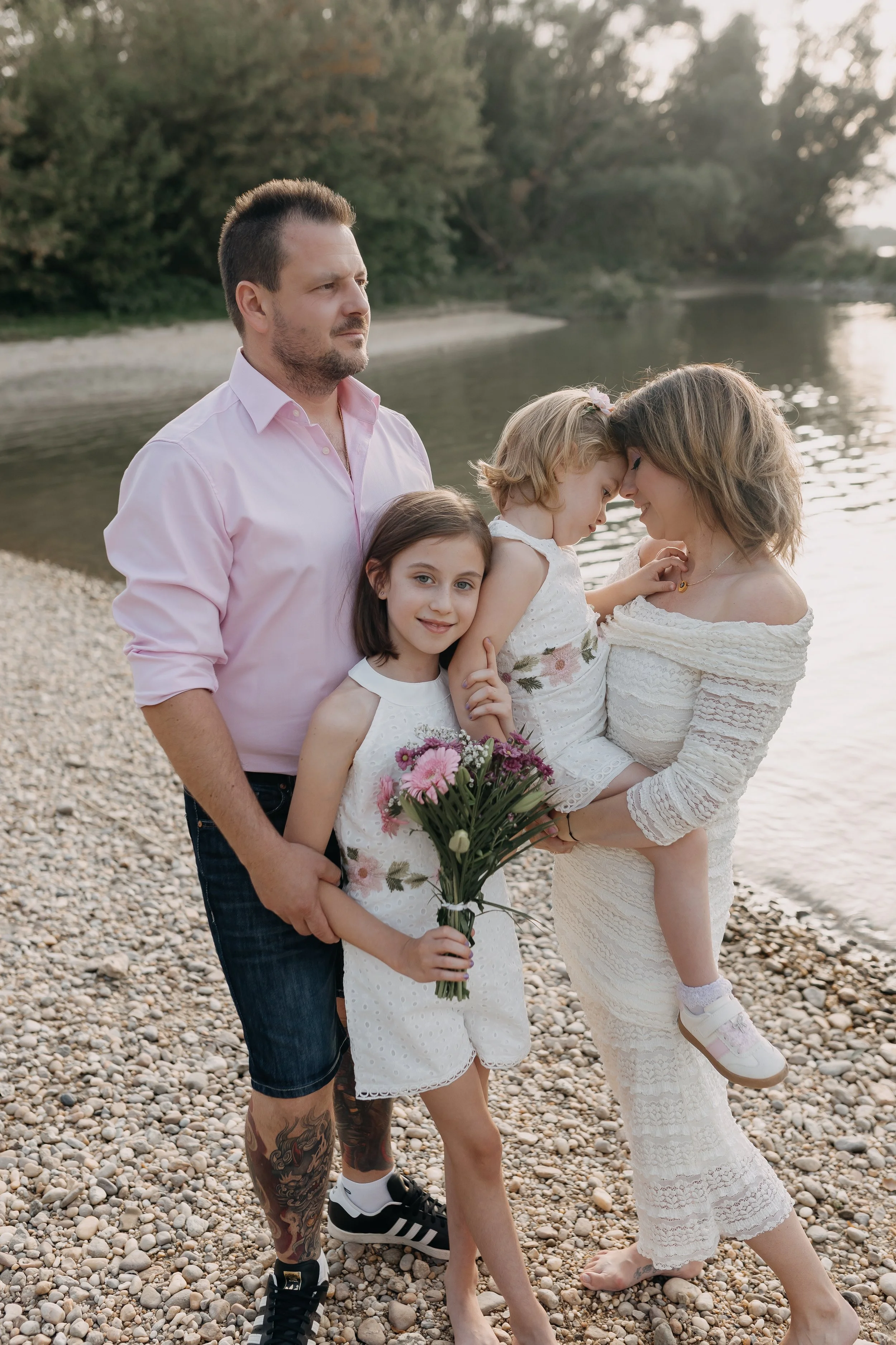 Family of five on a rocky beach by a river, with trees in the background. The father is in a pink shirt, the mother holds a young girl, and two other girls are in front, one holding a bouquet of flowers. They are all close together, touching forehead