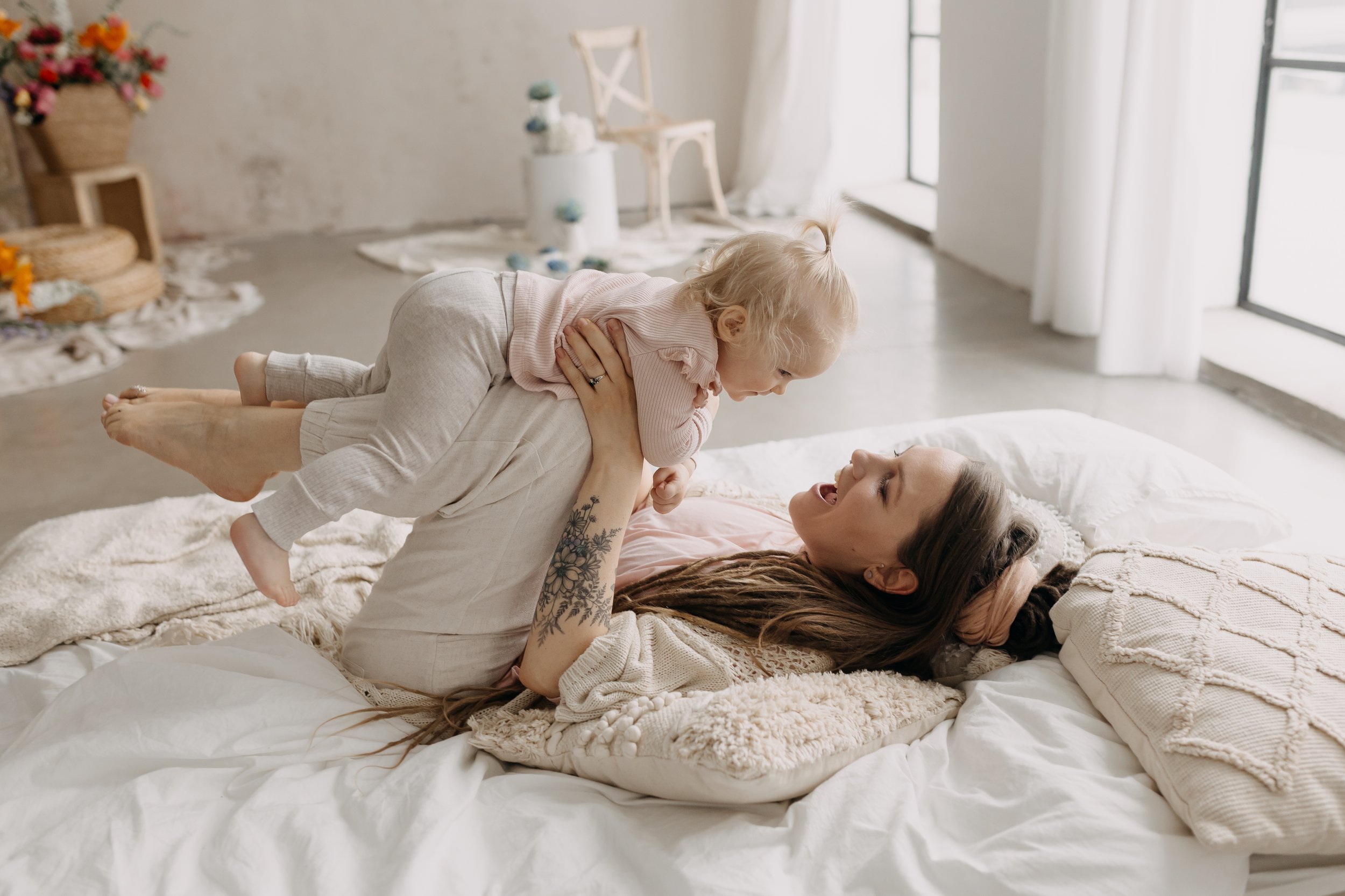 A woman lying on a bed holding a young child above her, both smiling and playing. The room is bright with natural light and decorated with flowers and soft furnishings.