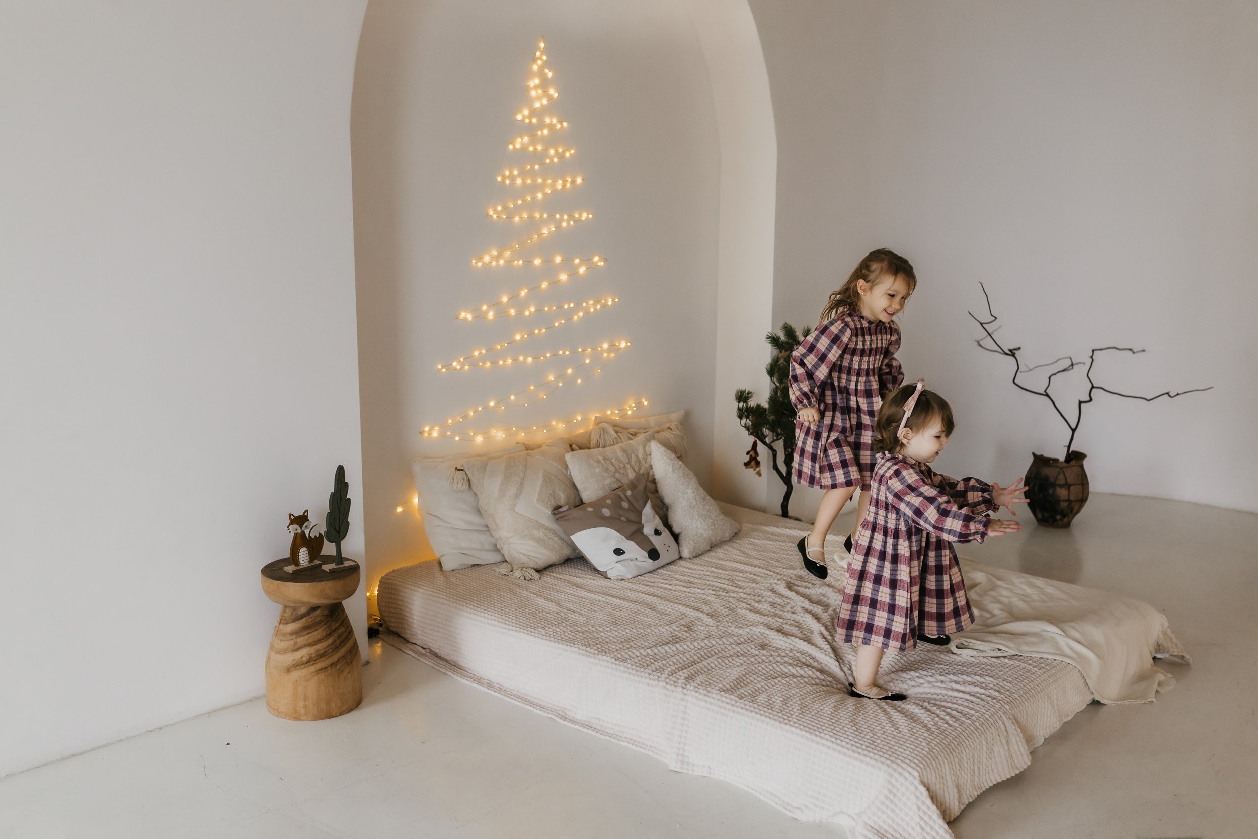 Two young girls in matching plaid dresses playing on a bed in a decorated room with a string lights Christmas tree on the wall and small holiday decor.