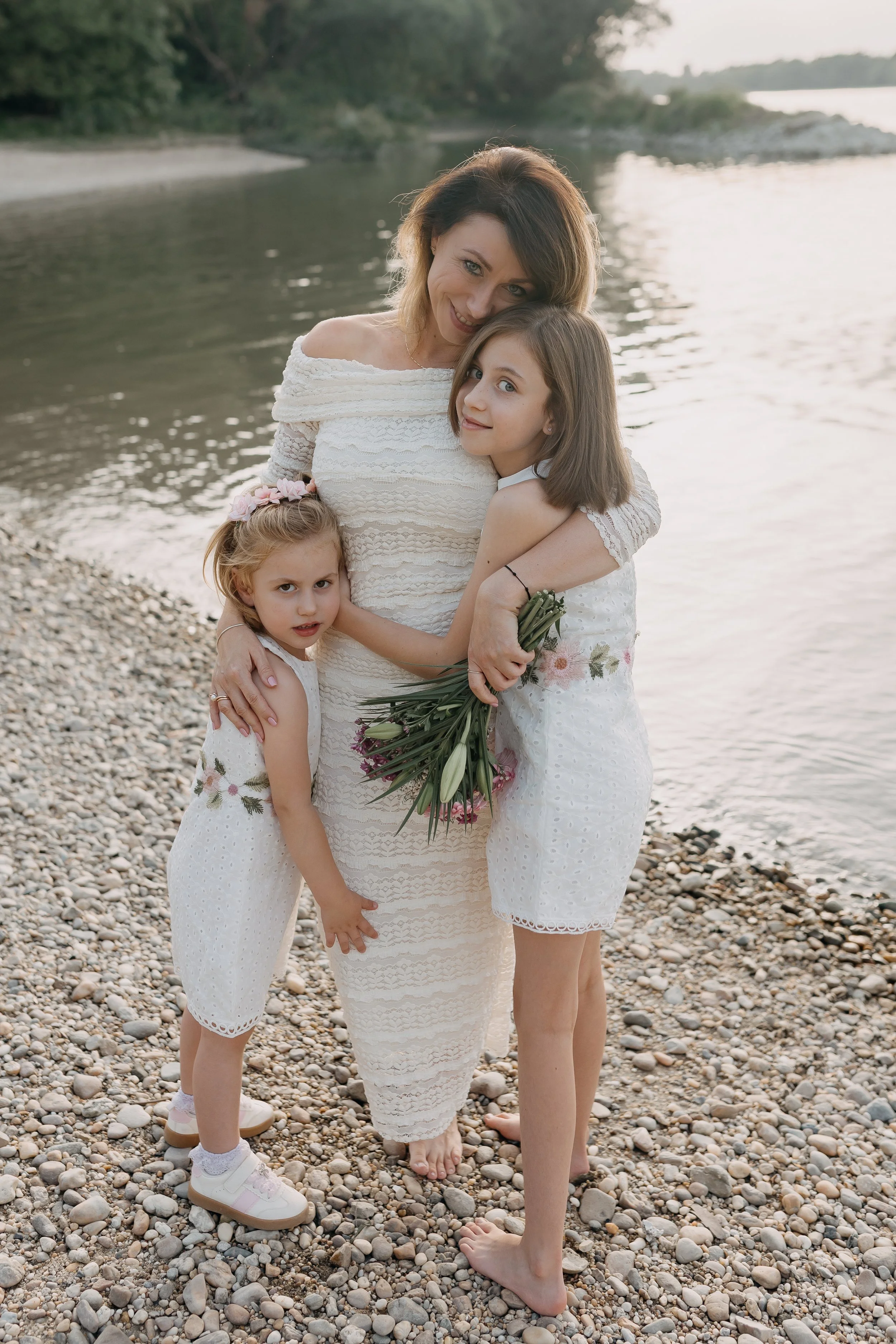 A woman and three young girls standing on a pebble beach near water, dressed in white, with the woman holding a bouquet of flowers, all smiling and hugging.